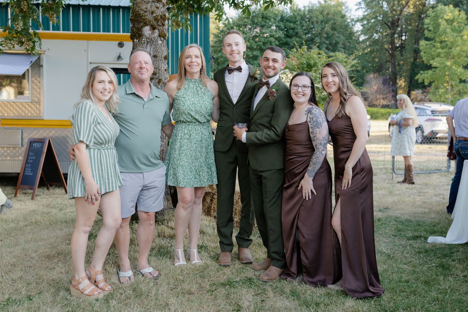 Wedding party group photo outdoors featuring two grooms in olive suits with bridesmaids in brown dresses and guests smiling.