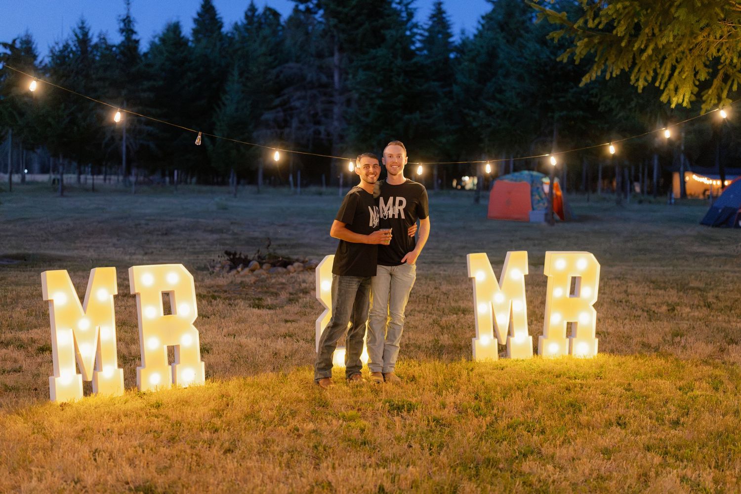 Couple posing between illuminated MR MR marquee letters at an outdoor evening wedding celebration.