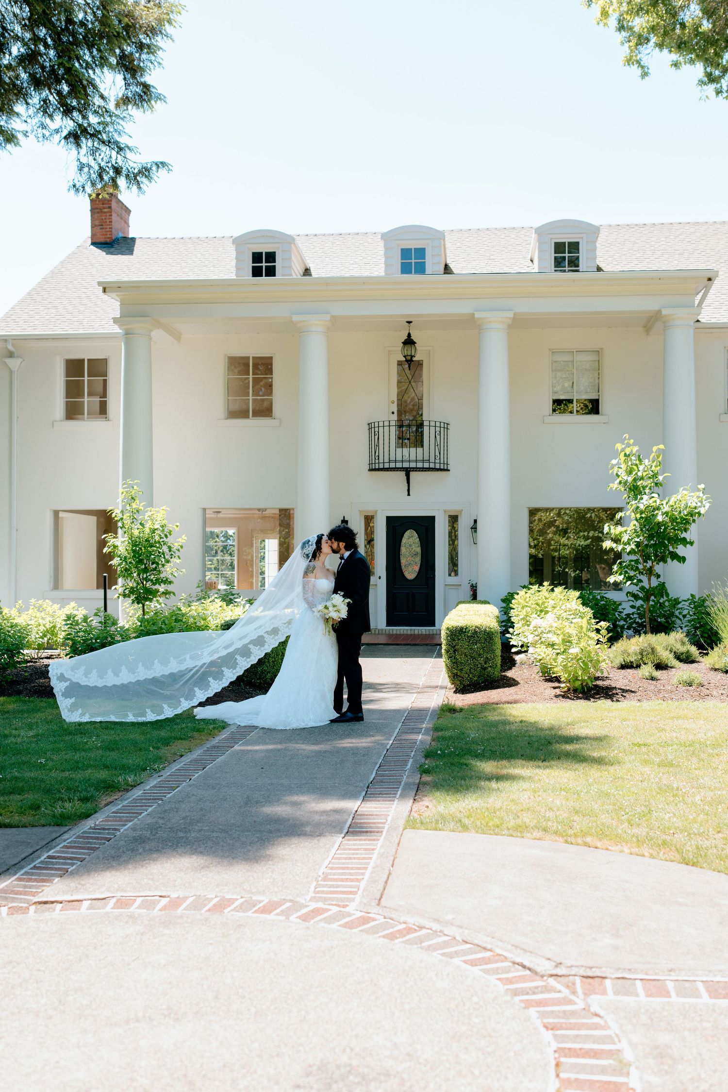 Bride and groom kissing in front of elegant white colonial mansion with tall columns, bride's veil flowing in the breeze.