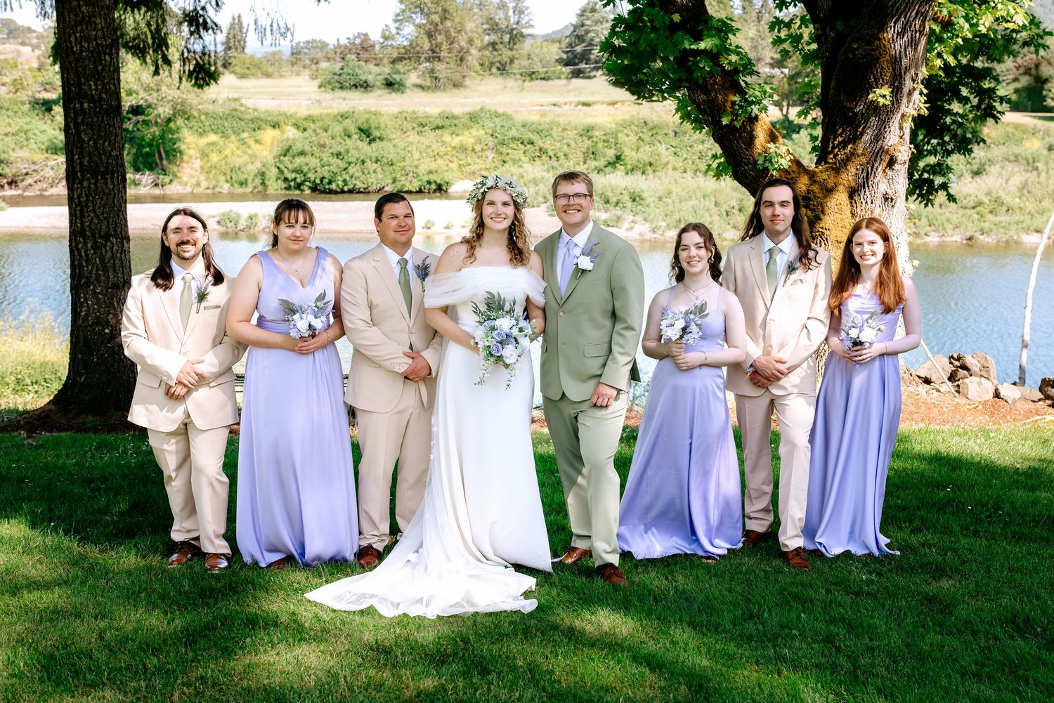 Wedding party posing outdoors with bride and groom flanked by bridesmaids in lavender dresses and groomsmen in tan suits by a river.