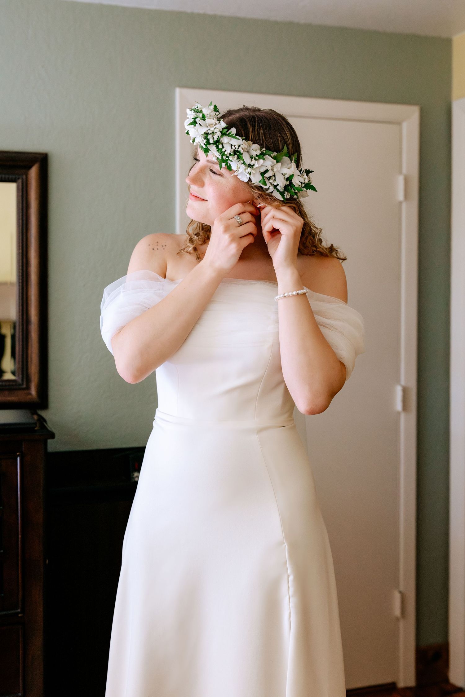 Bride in white off-shoulder gown adjusting floral crown with white flowers in her hair before wedding.