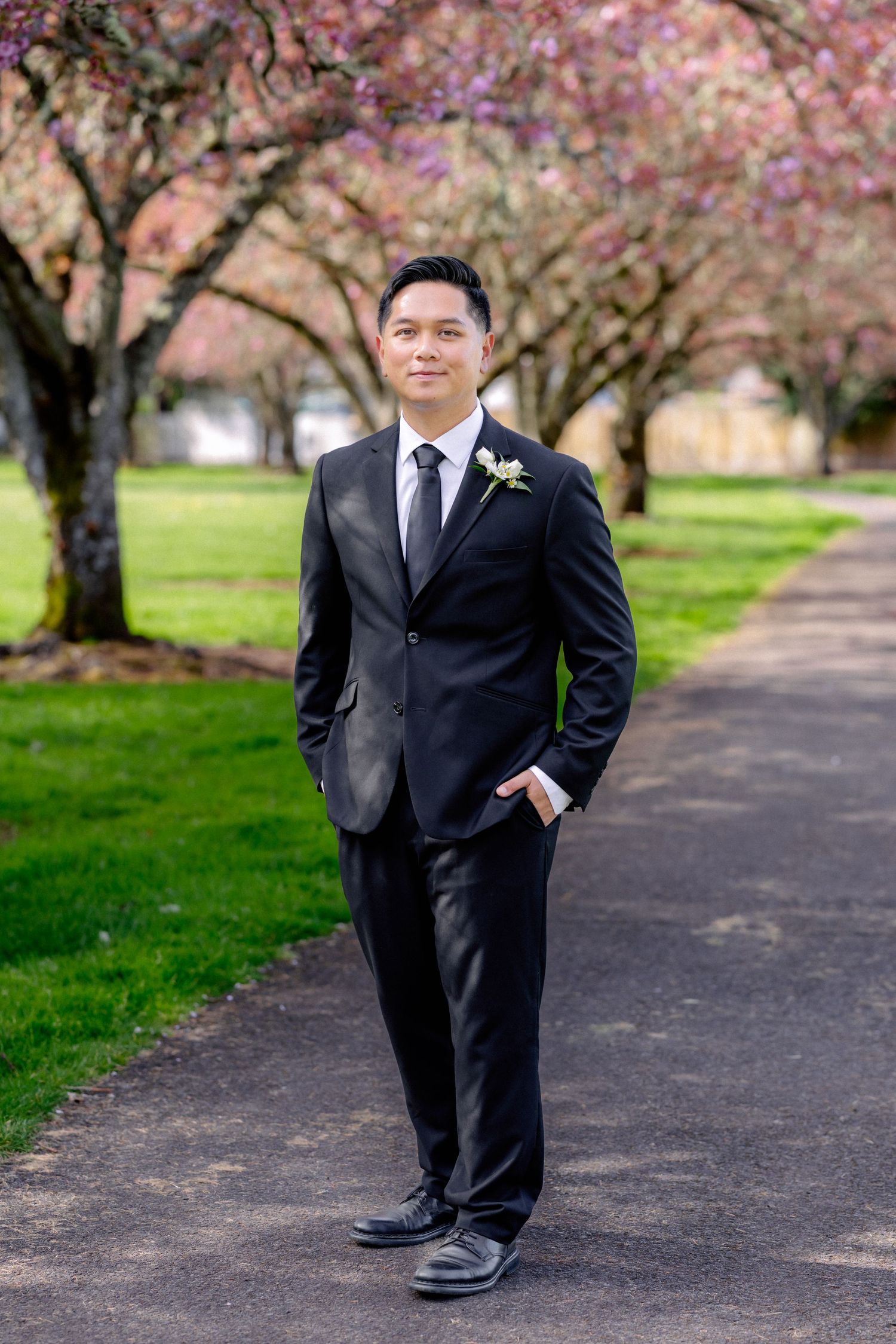 Groom in black suit with boutonniere stands on park path lined with pink cherry blossom trees.