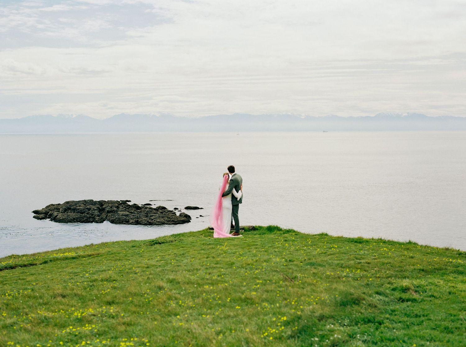 Couple embracing on a grassy cliff overlooking a calm ocean on their wedding day.