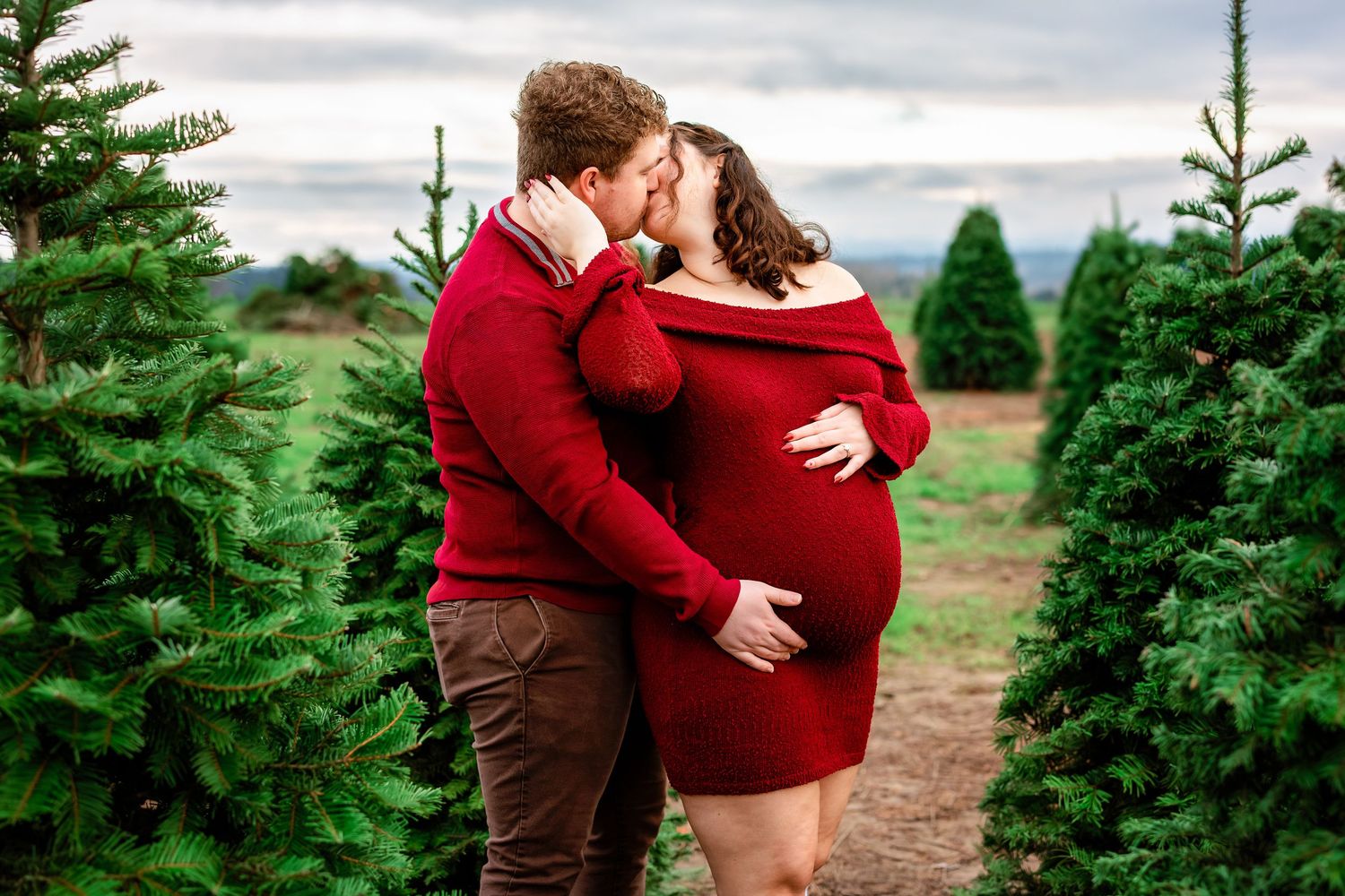 A couple in red and brown clothing embraces among evergreen trees at a Christmas tree farm during sunset.