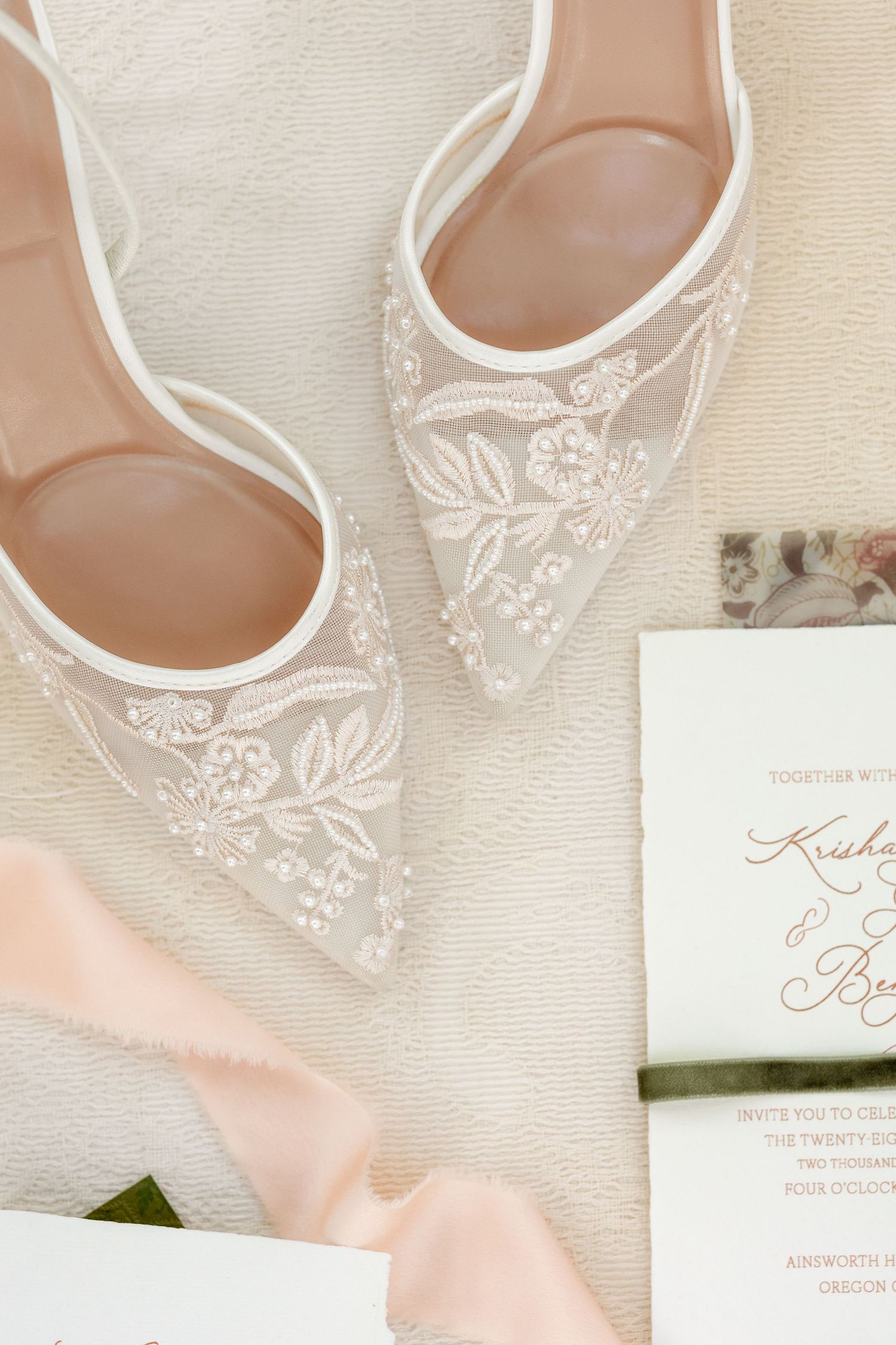 Delicate lace wedding flats with floral embroidery detail alongside invitation on cream background.