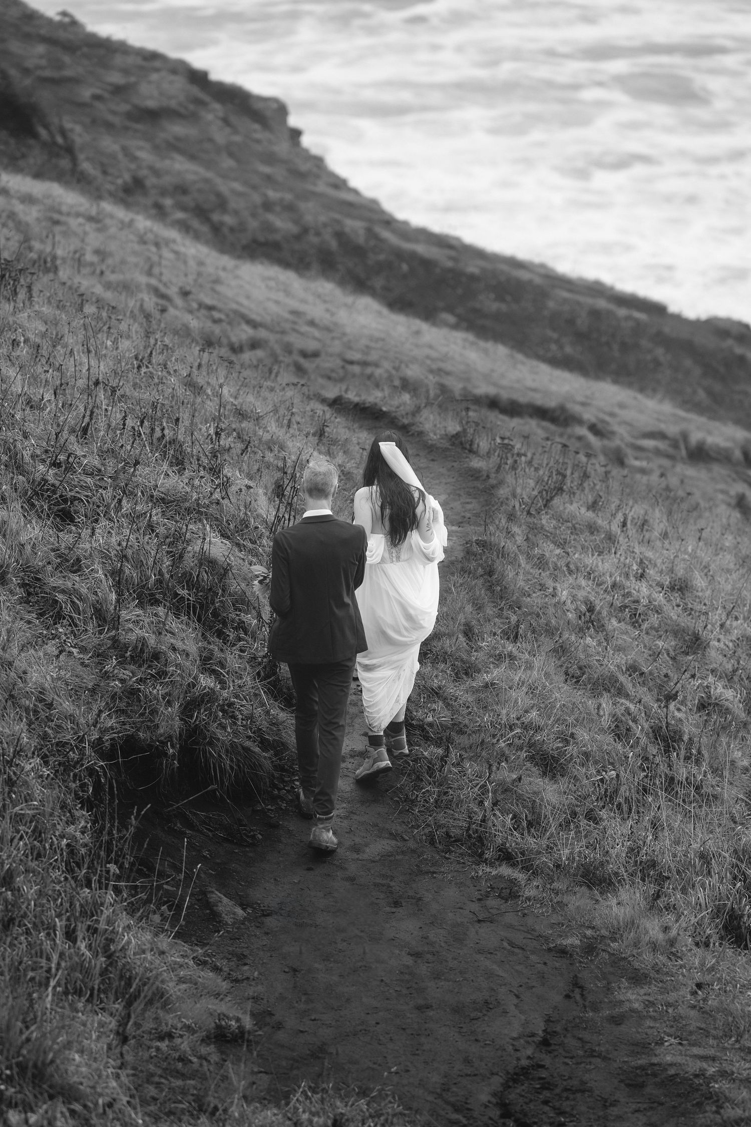 A couple in wedding attire walks up a rugged mountain path in black and white photography.