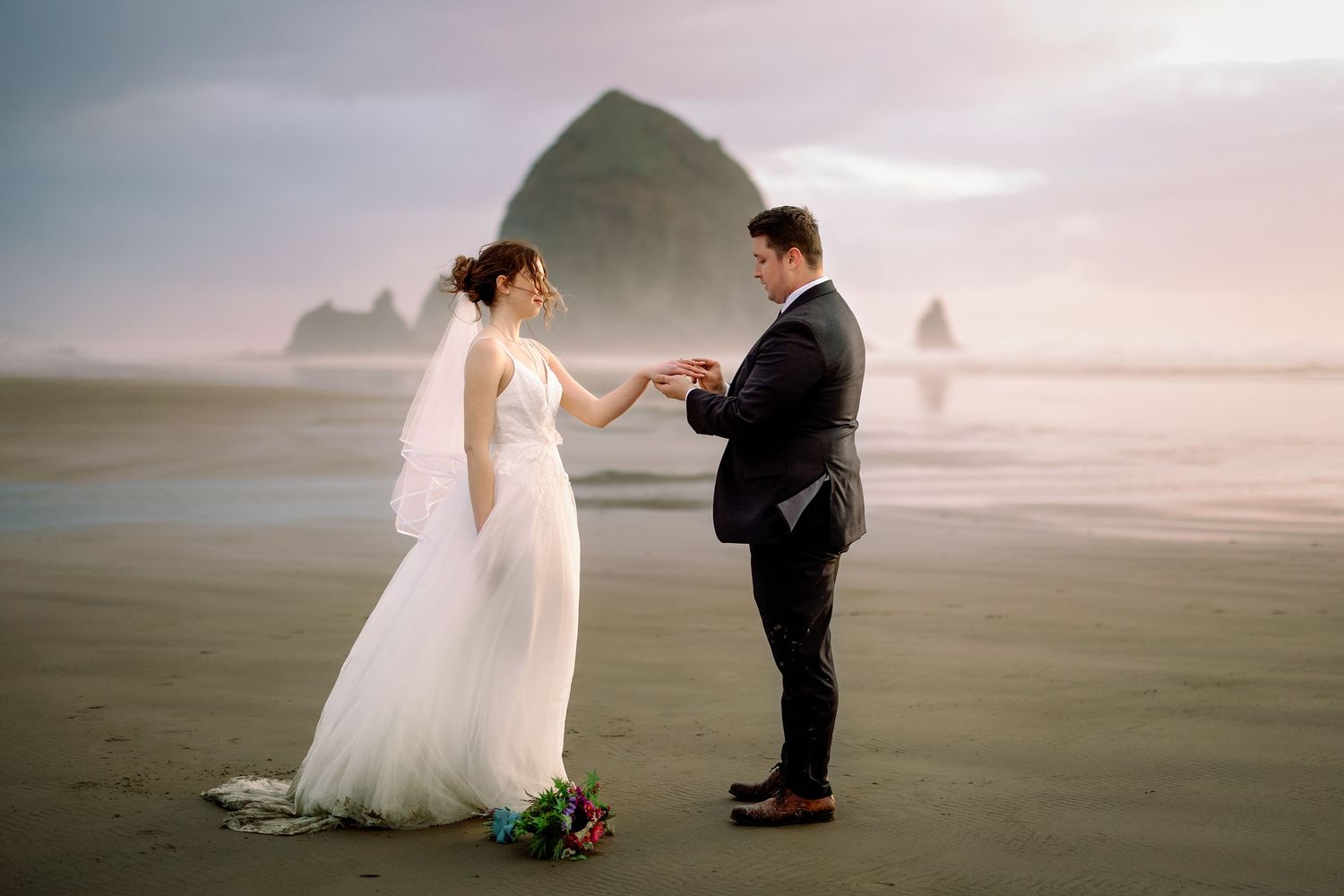 A romantic beach wedding scene with coastal rock formations and misty atmosphere during sunset at Cannon Beach, Oregon.