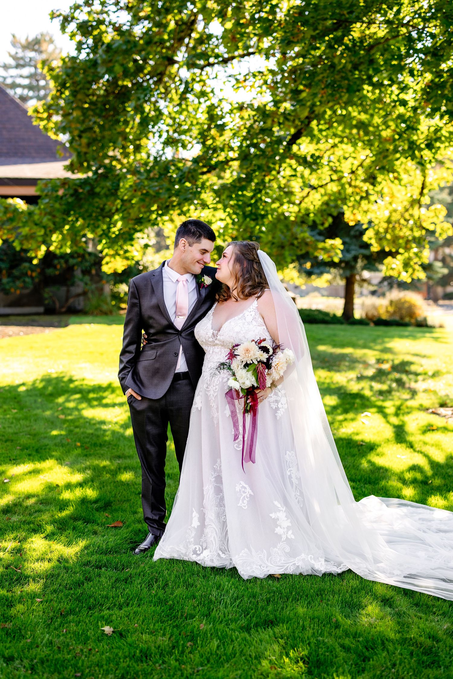 Newlyweds share an intimate moment on a sunlit lawn with beautiful trees in the background.