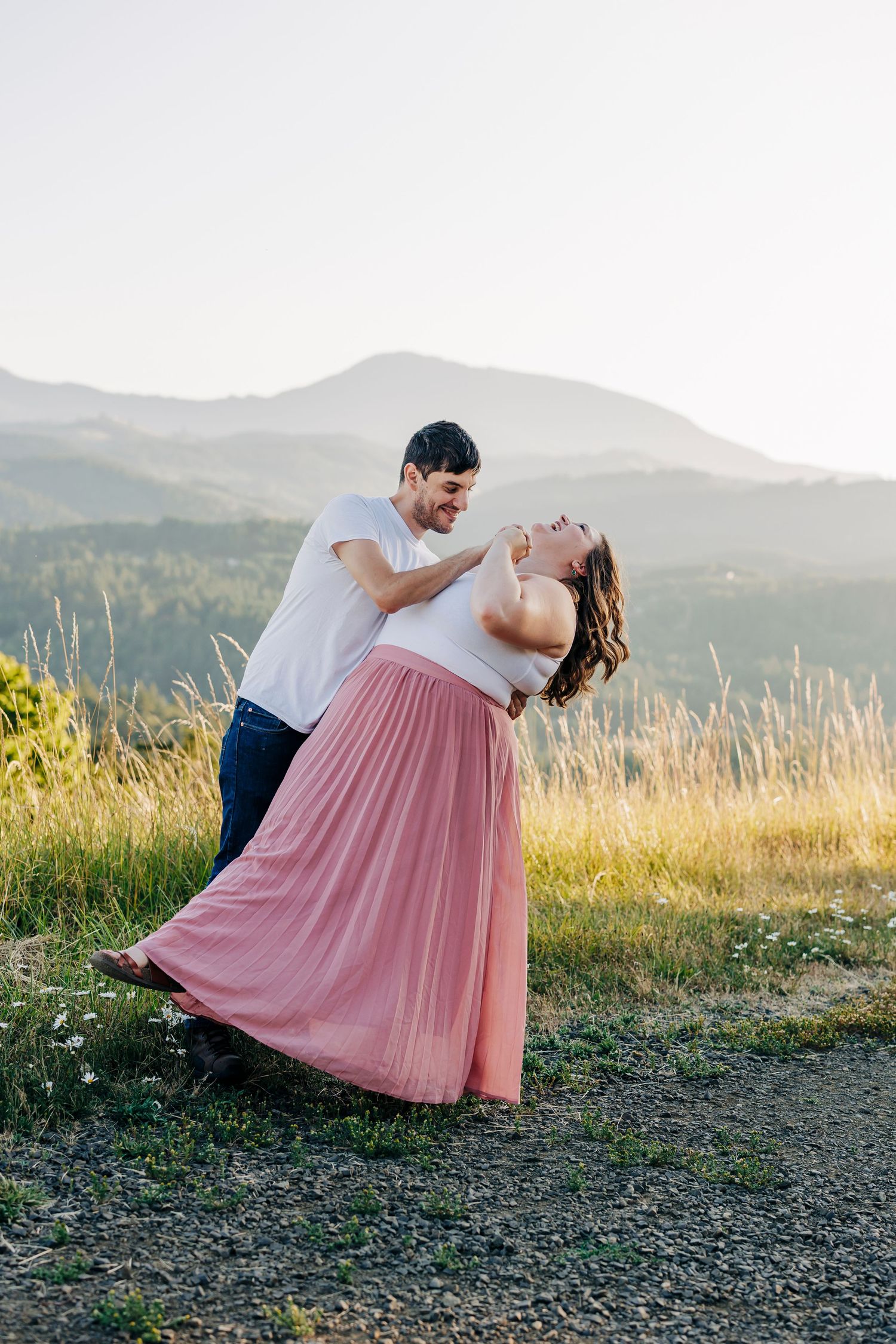 A couple shares a romantic moment against a mountain backdrop as the sunlight illuminates a golden field.