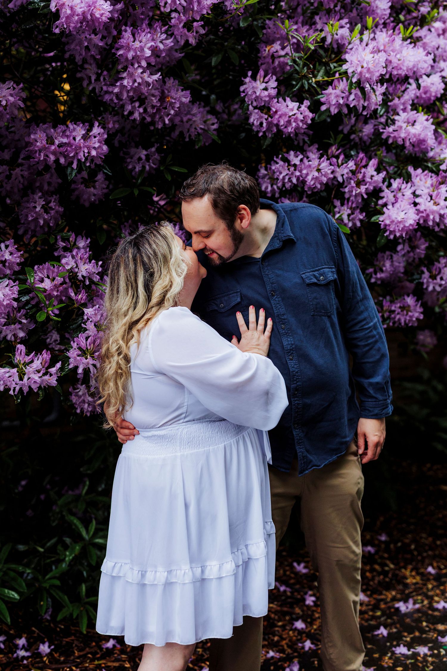 A couple shares an intimate moment in front of blooming purple rhododendrons in a spring garden.