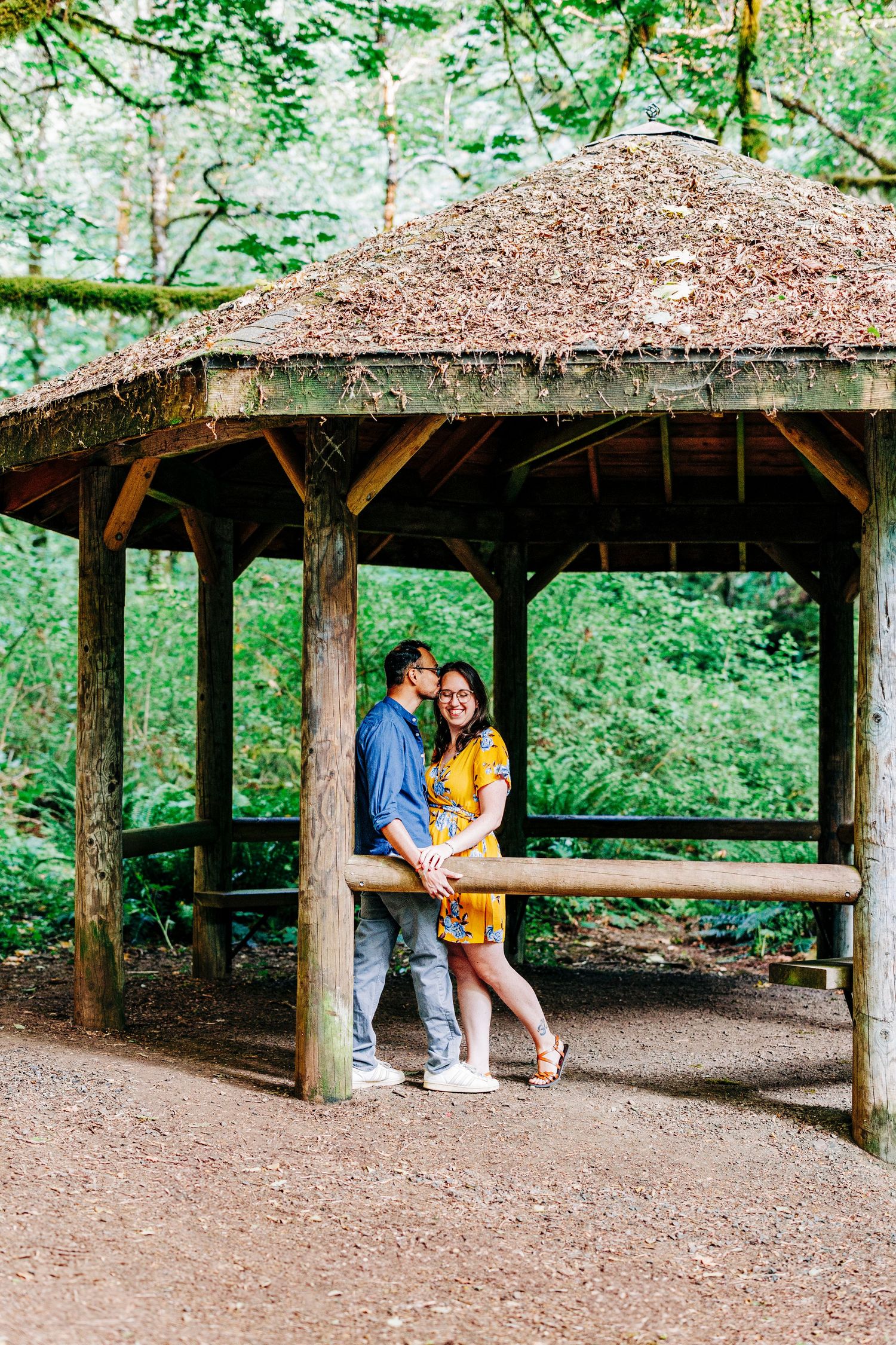 A couple embraces under a rustic wooden gazebo with a thatched roof surrounded by lush green forest.