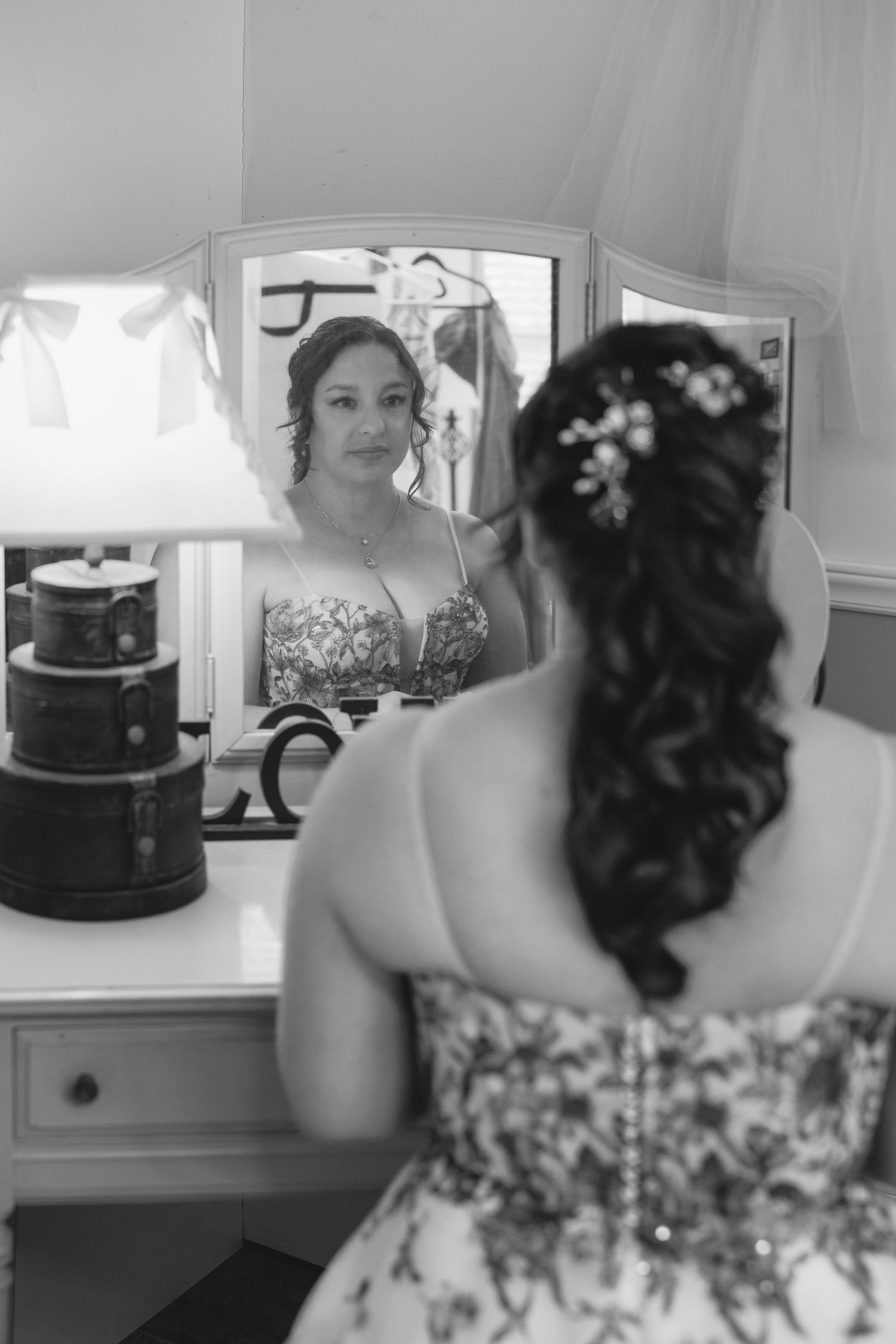 Black and white photo showing preparation scene with floral dress details and mirrors.