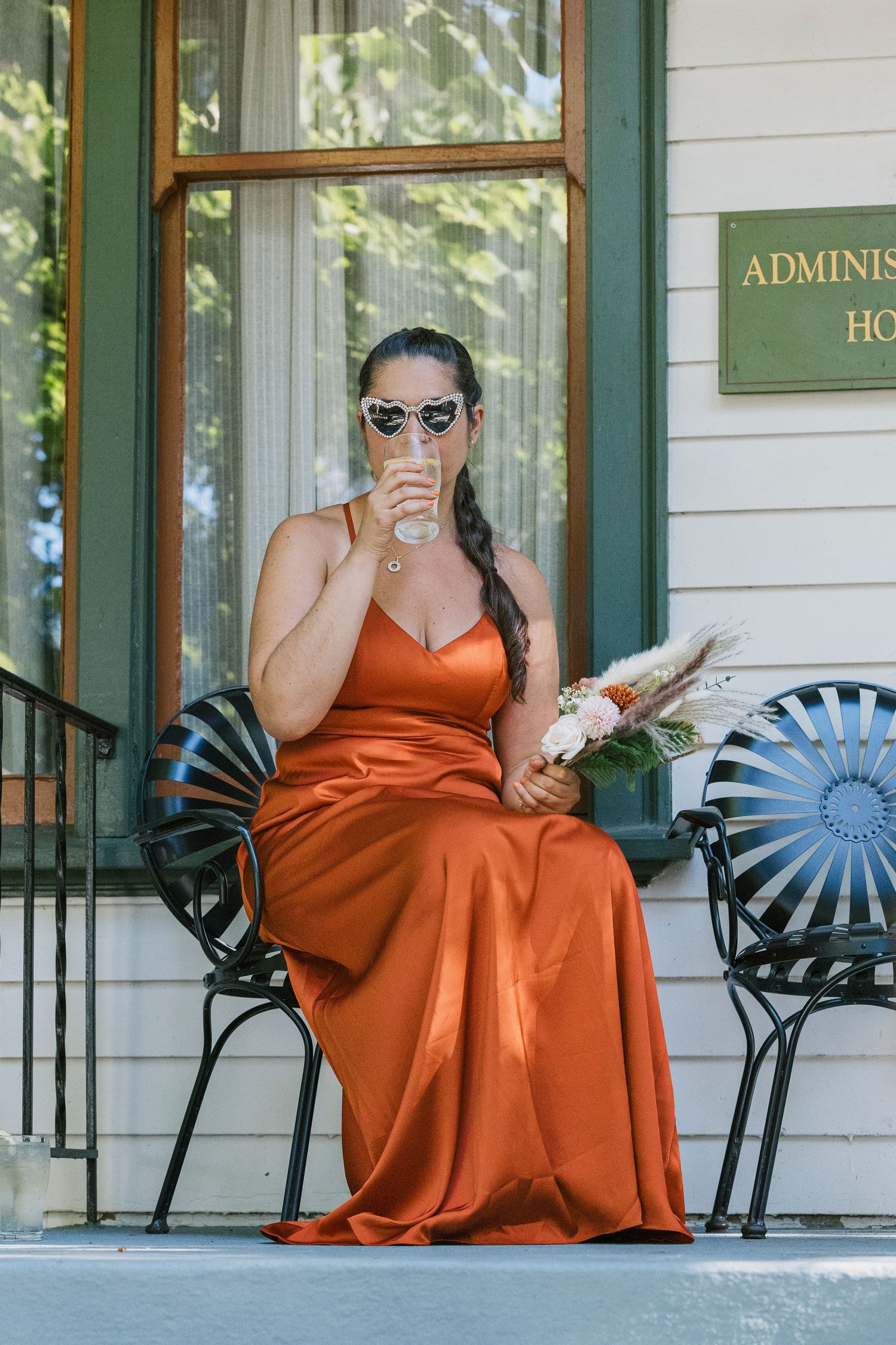 A person wearing an orange dress relaxes in a black metal chair outside a white building with green trim.