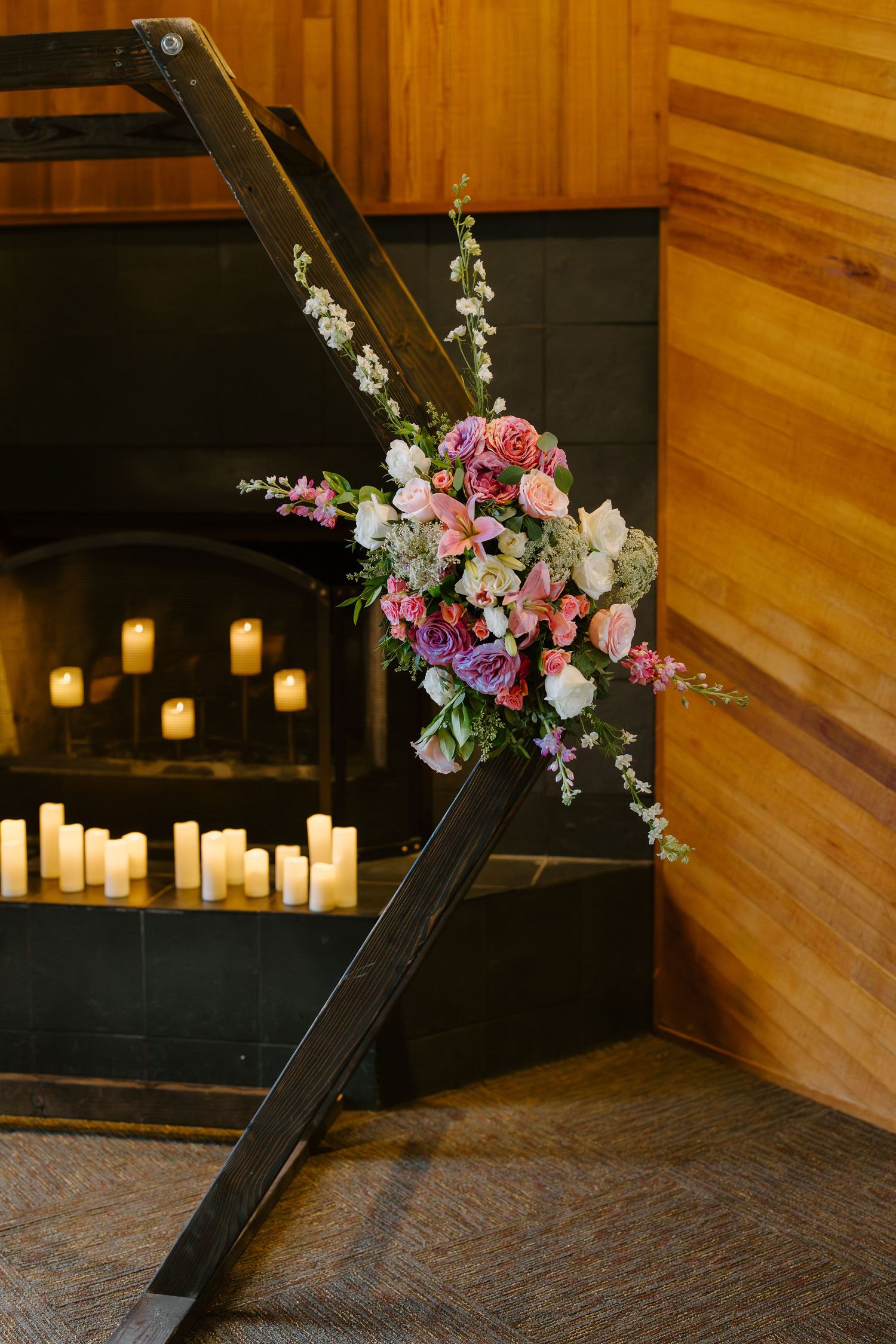 Pink and purple floral arrangement on wooden aisle marker beside candlelit fireplace at wedding.