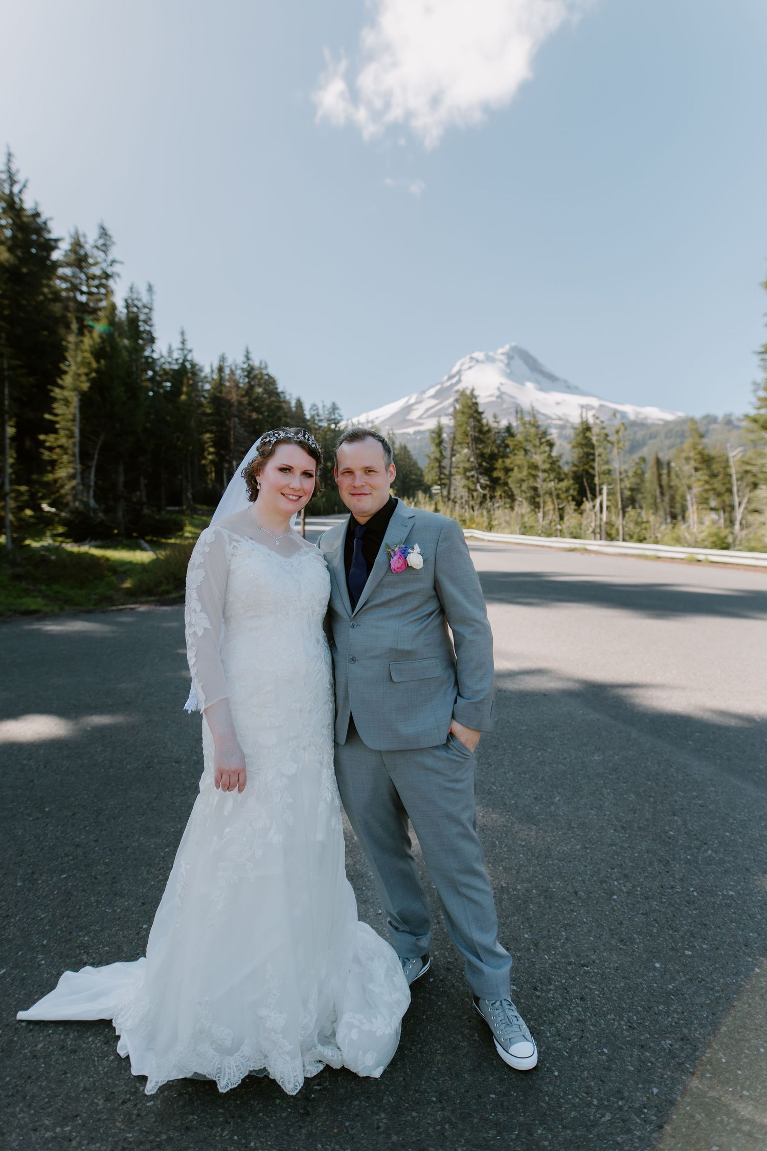 Bride and groom pose on mountain road with snow-capped Mt. Hood and evergreen trees in background on wedding day.