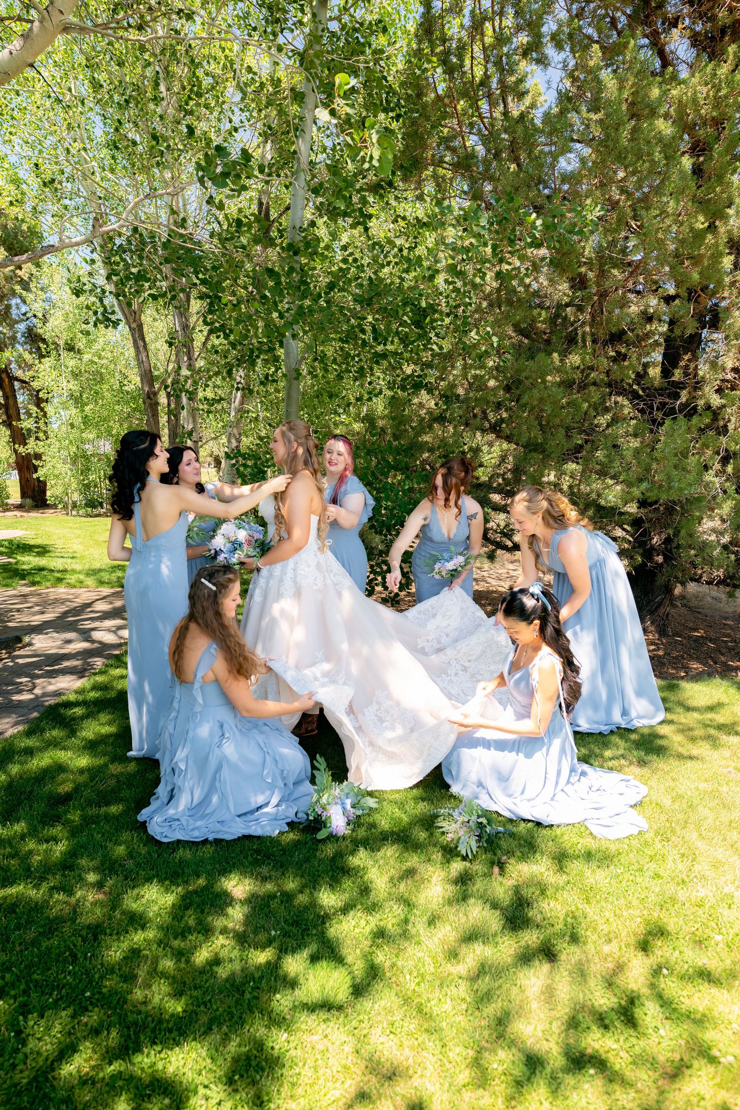 Bride in white gown surrounded by bridesmaids in blue dresses on green lawn.