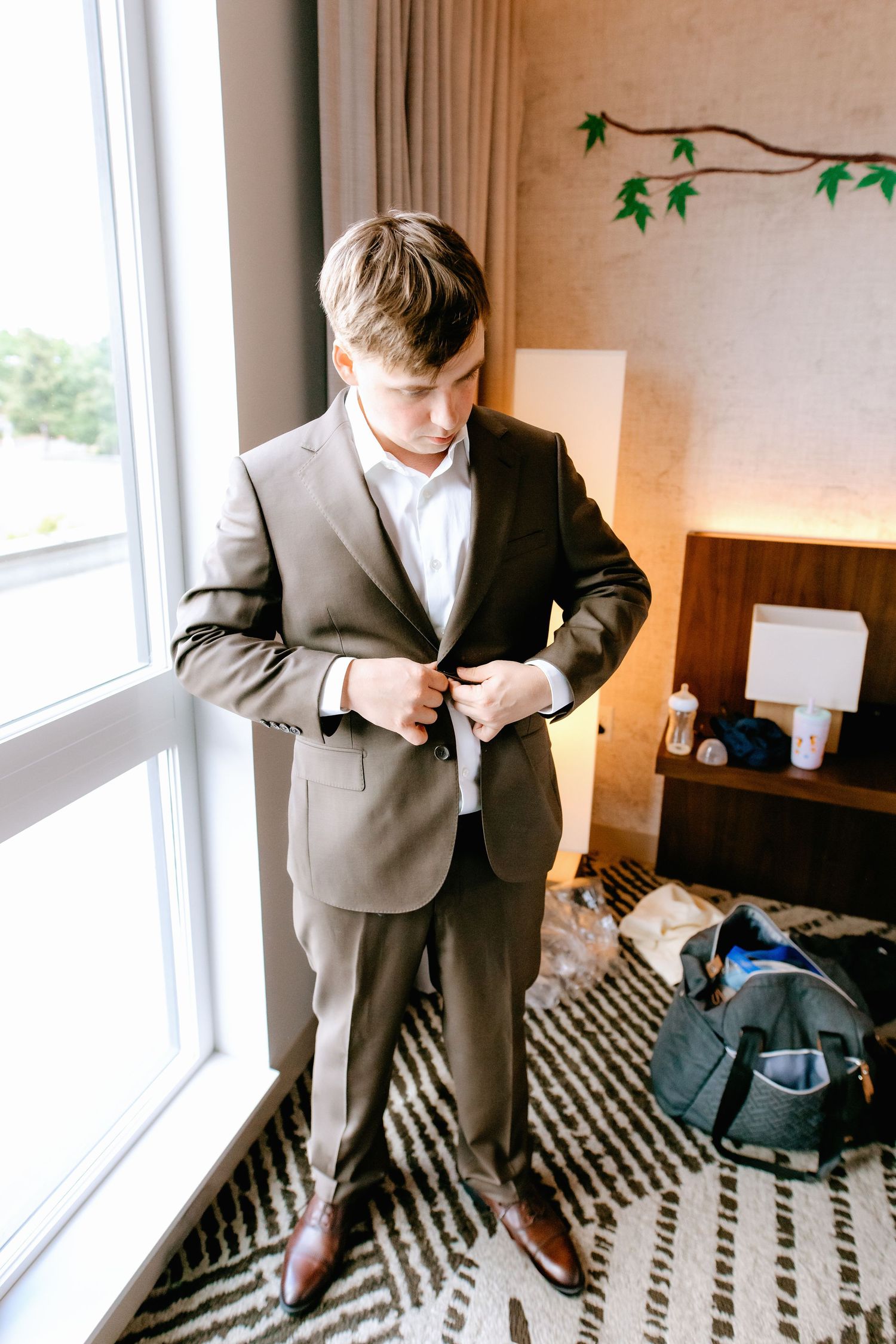 Groom buttoning his gray suit jacket while standing near a large window in a hotel room before his wedding.