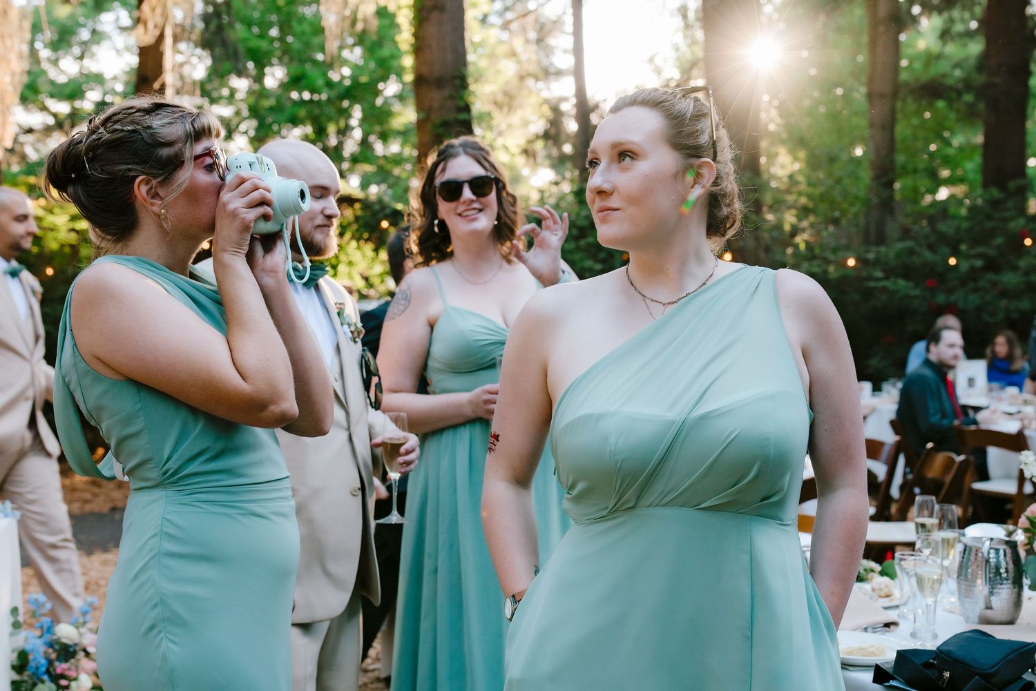 Bridesmaids in sage green gowns gather outdoors at a woodland wedding reception, with sunlight filtering through tall trees.