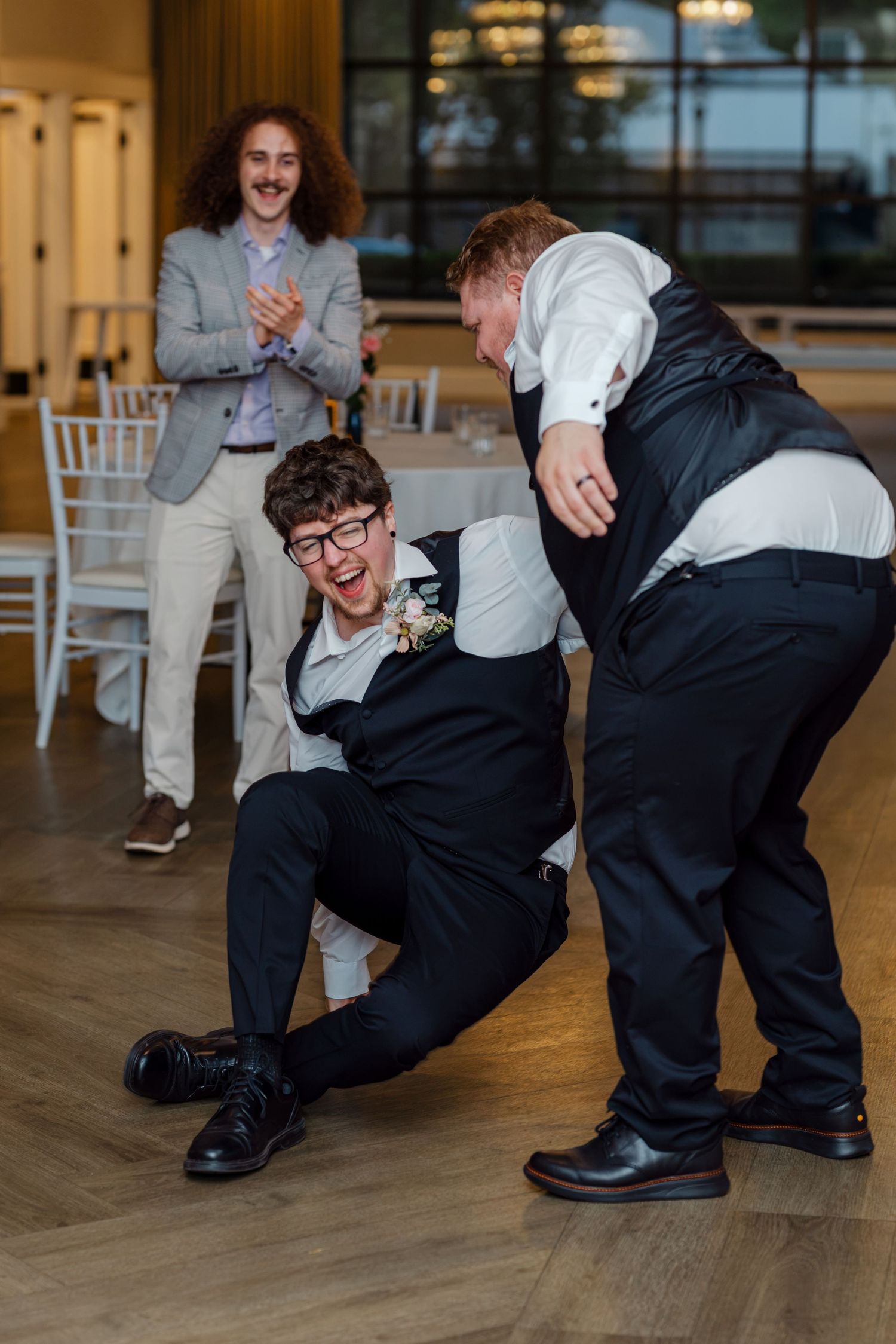 Groomsmen laughing and playing on dance floor at wedding reception with guests watching.
