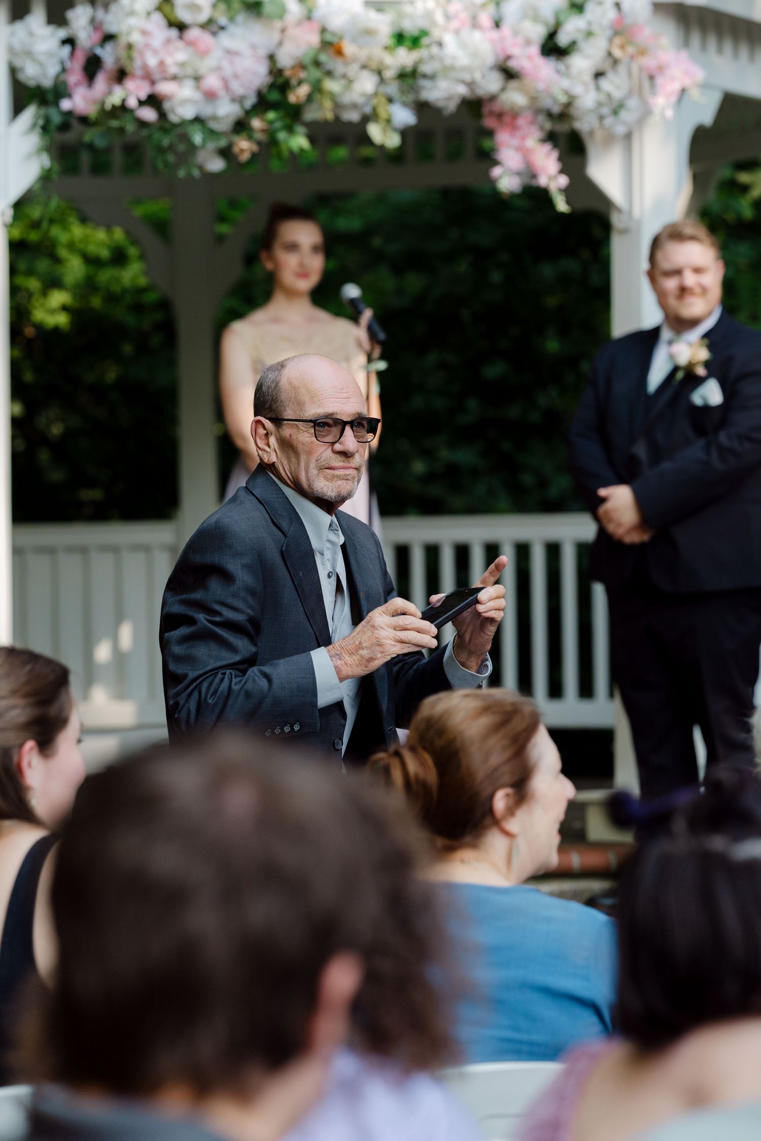 Elderly man in suit speaking at outdoor wedding ceremony near floral gazebo with guests seated.