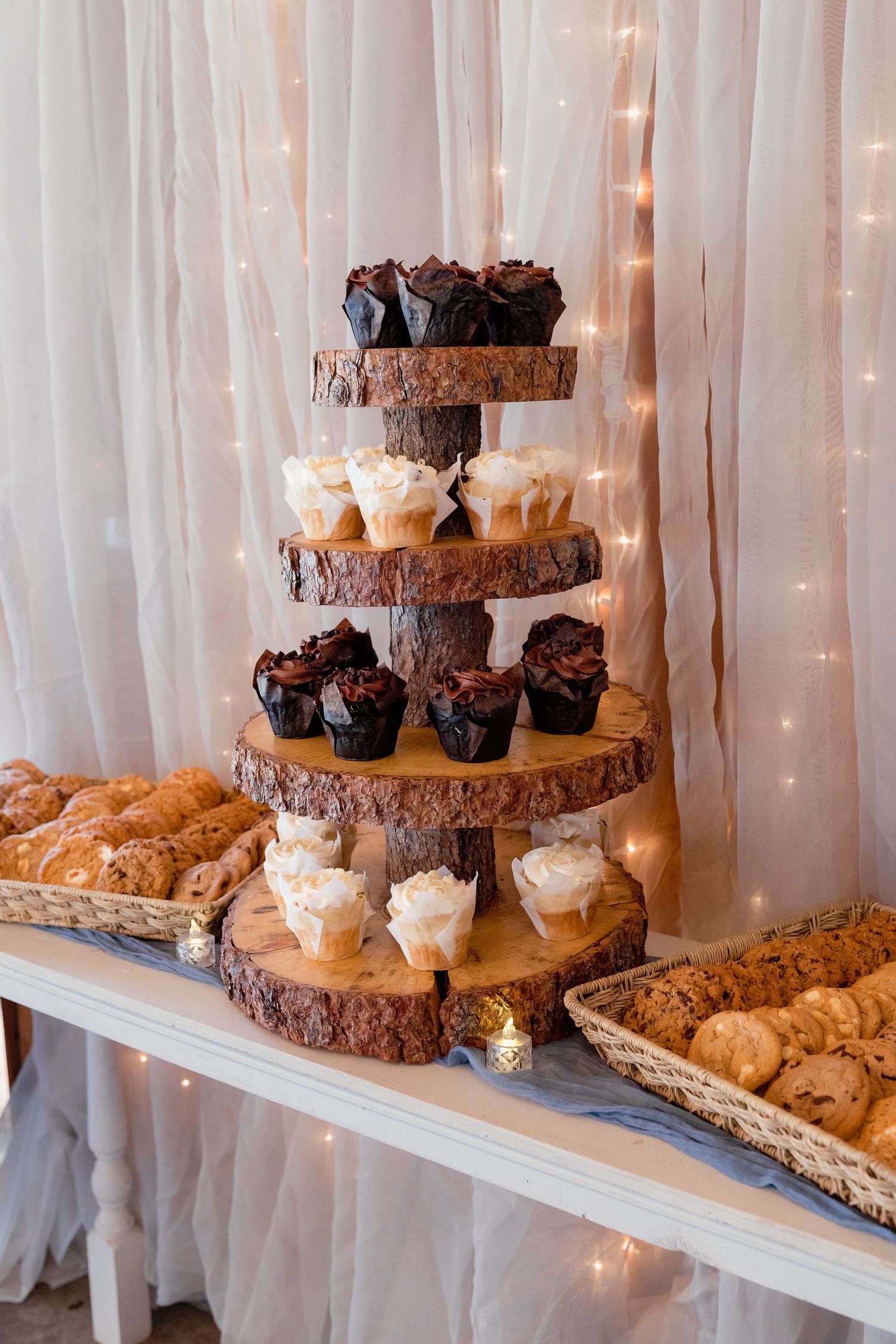 Elegant rustic wedding dessert display with tiered wood slab stand holding cupcakes surrounded by cookies and pastries.