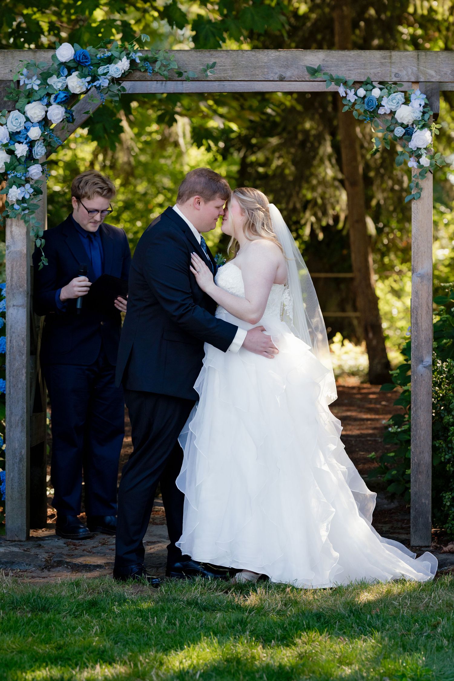 Bride and groom sharing first kiss under rustic wooden arch decorated with blue and white flowers.