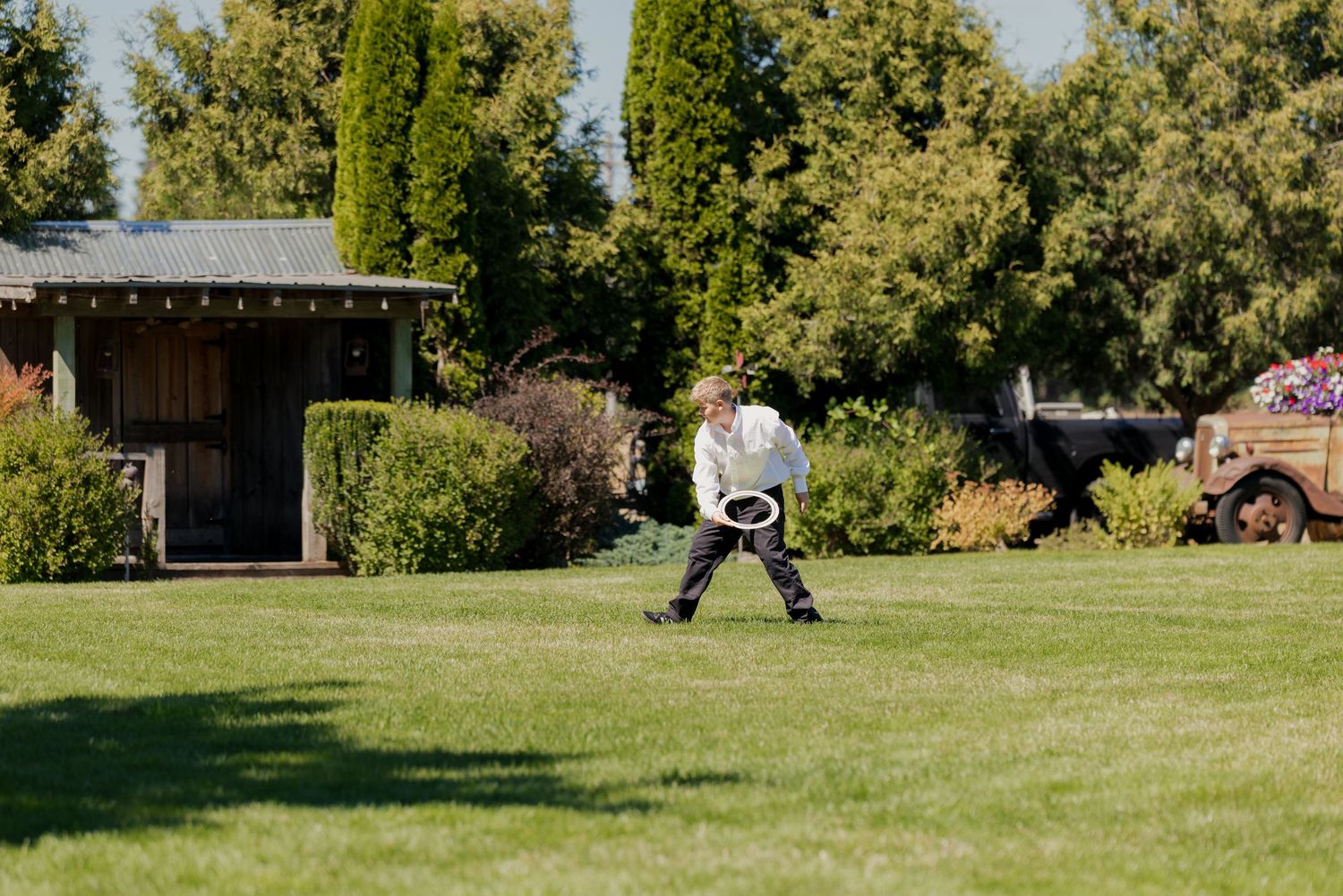 Person playing frisbee on lush green lawn surrounded by trees and rustic garden buildings on sunny day.