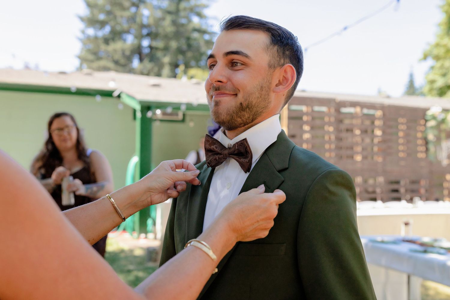 Groom in green suit getting bow tie adjusted before wedding ceremony outdoors.
