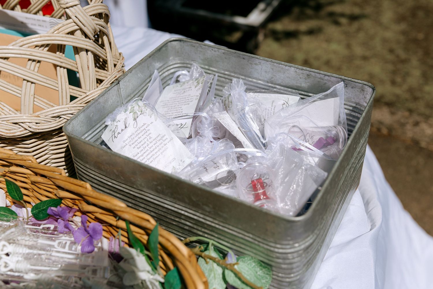 Metal tin filled with wrapped wedding favor items displayed on a floral tablecloth beside a wicker basket.