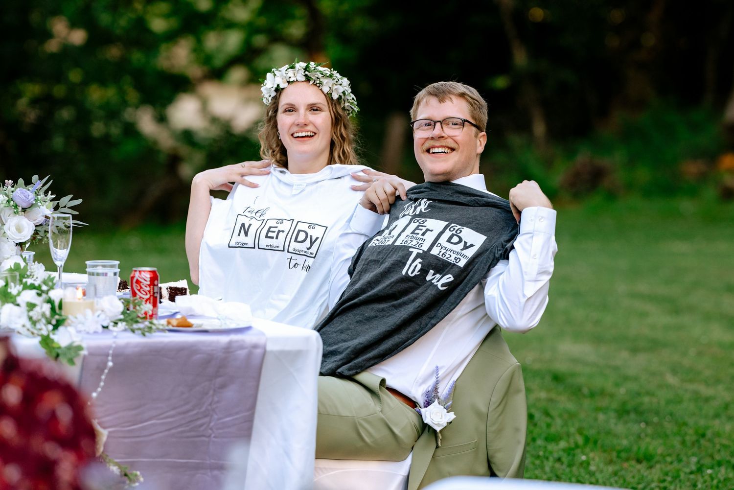 Smiling newlyweds hold up matching nerdy periodic table t-shirts at outdoor wedding reception.