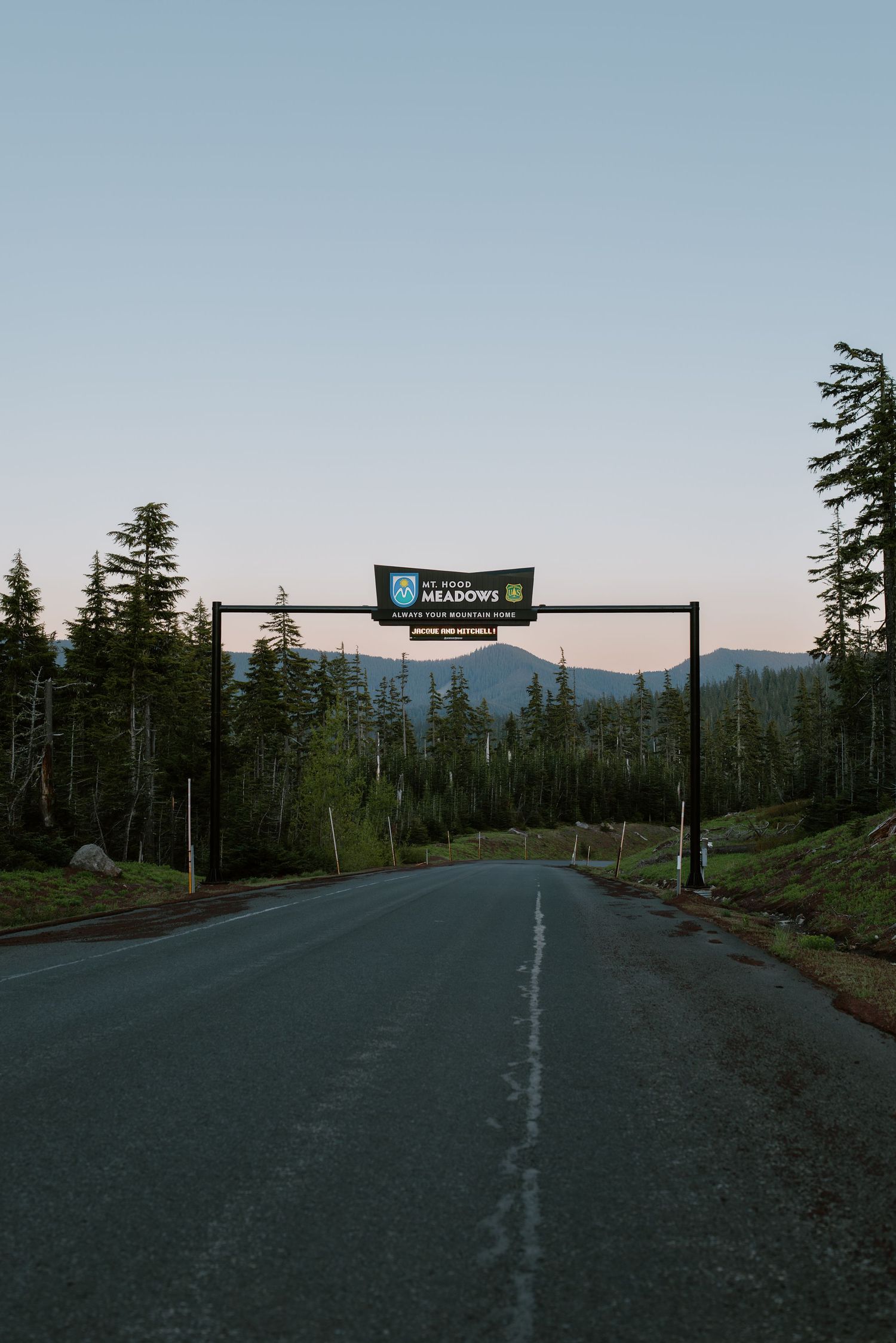 Road leading to Mt. Hood Meadows ski resort entrance sign surrounded by evergreen trees at dusk.