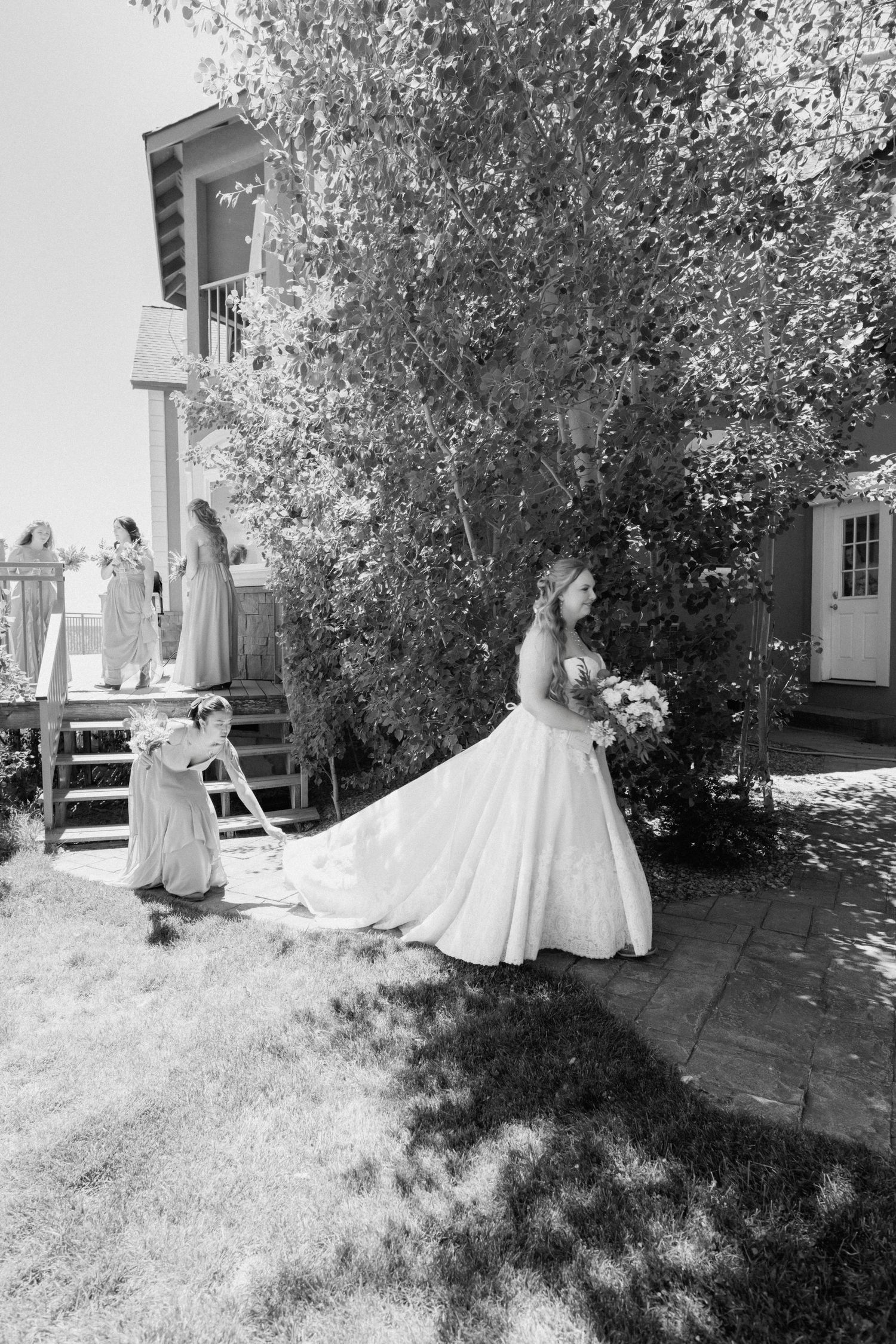 Bride in elegant white gown with long train stands outdoors with flower girl in black and white photo.