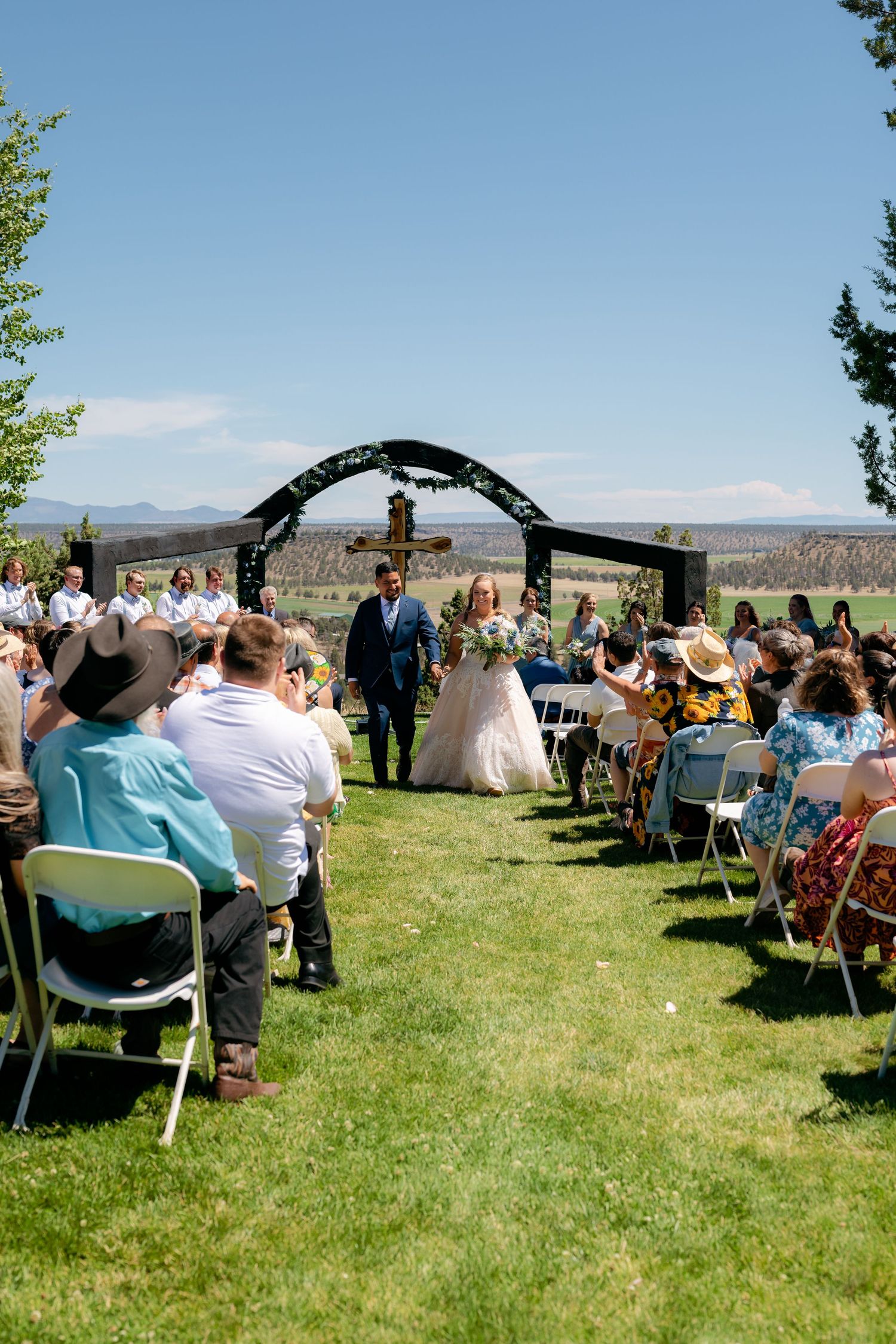 Bride walks down outdoor aisle with guests seated on green lawn under blue sky.