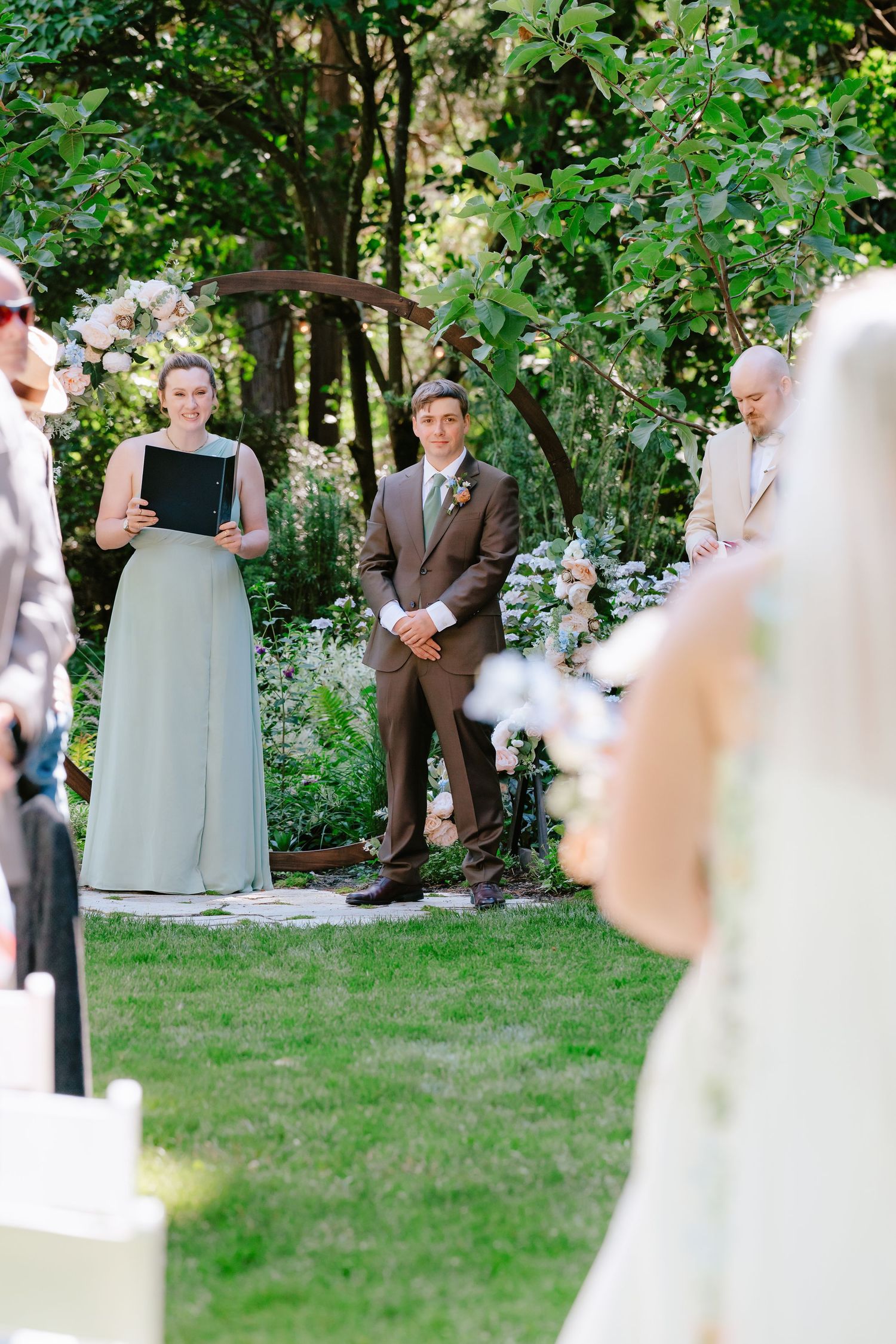 Groom in brown suit watches bride walk down aisle at outdoor garden wedding ceremony with floral arch.