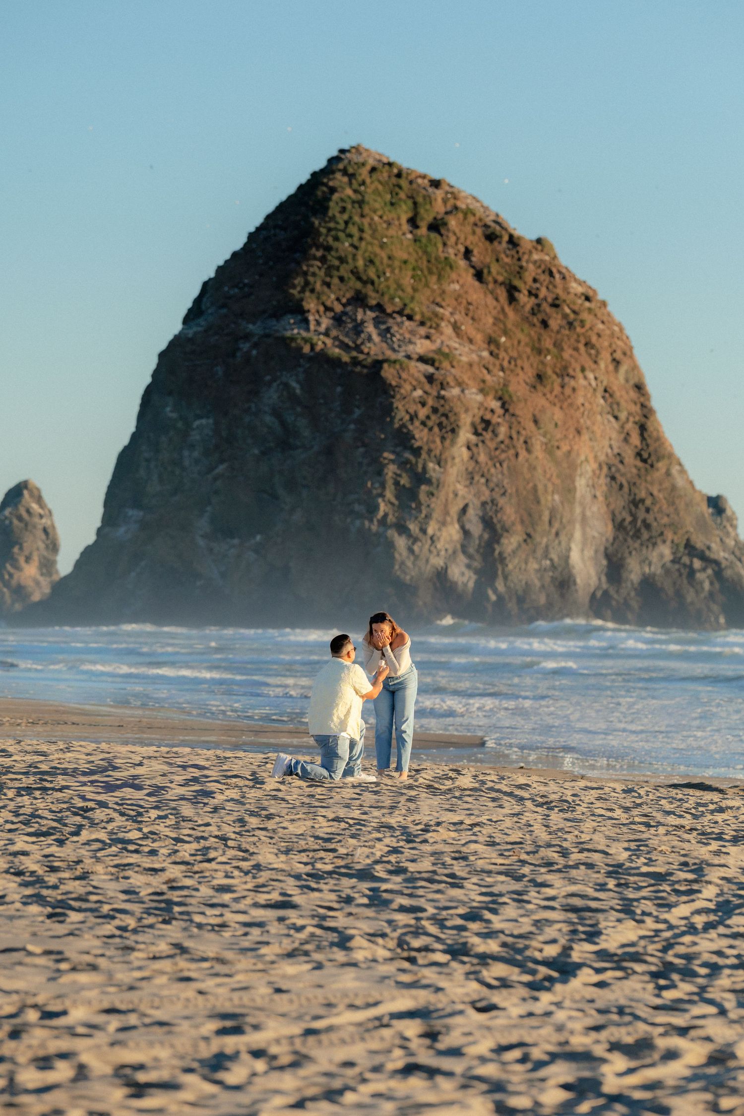 Couple sharing a marriage proposal on the sandy beach at Cannon Beach with iconic Haystack Rock in the background.