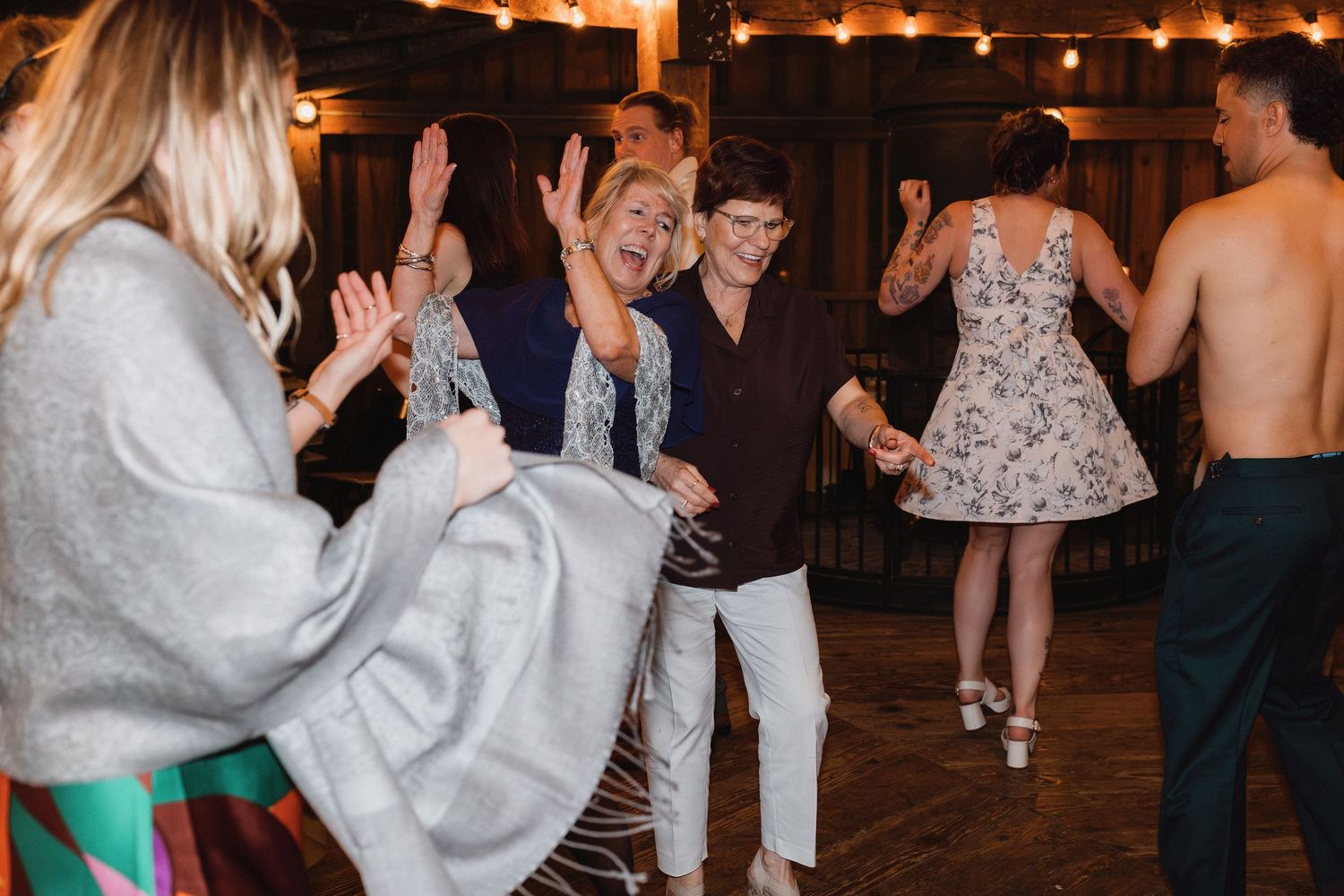 Joyful wedding guests dancing and celebrating together under warm string lights at indoor reception.