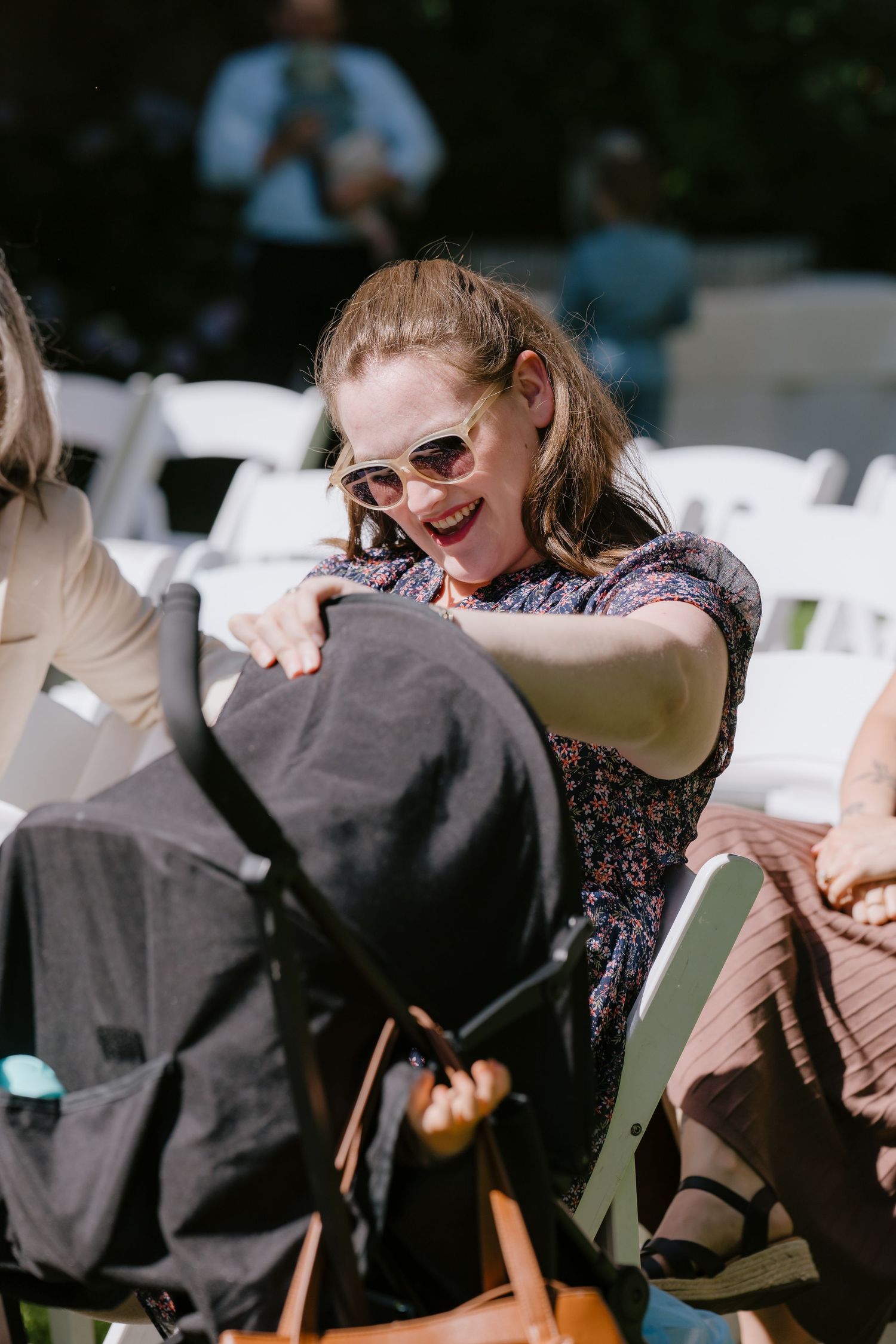 A smiling woman in sunglasses reaches toward a black stroller at an outdoor wedding ceremony with white chairs.