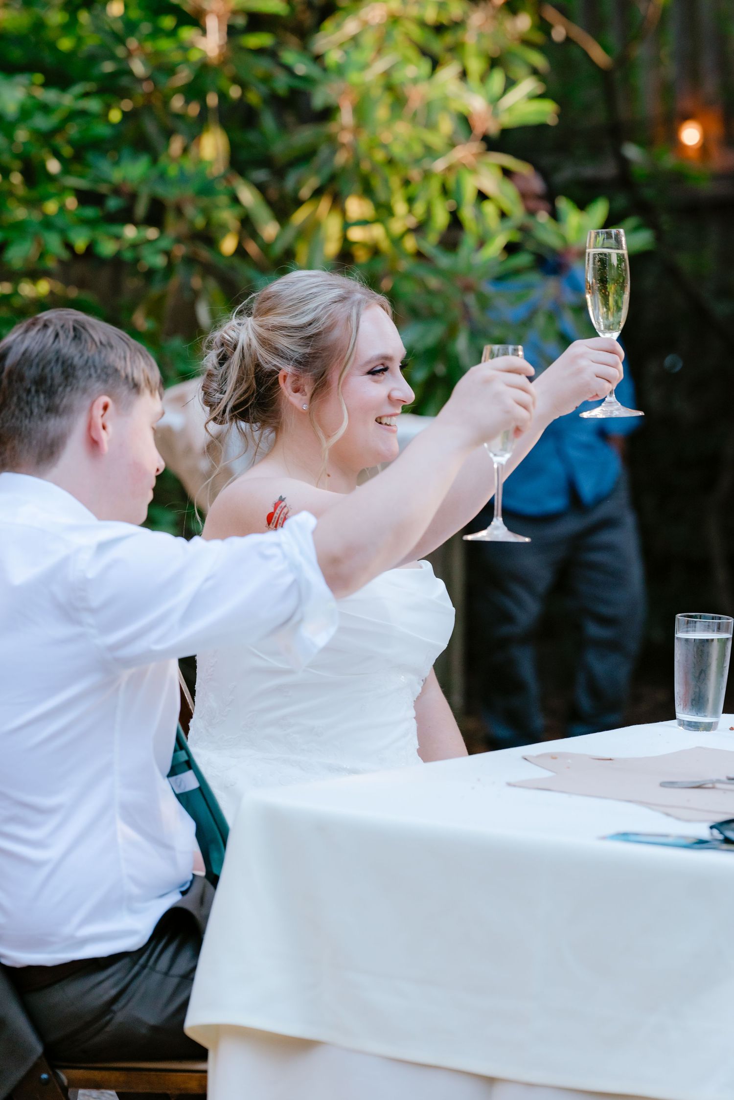 Smiling bride in white dress raising a champagne glass toast at outdoor wedding reception dinner table.