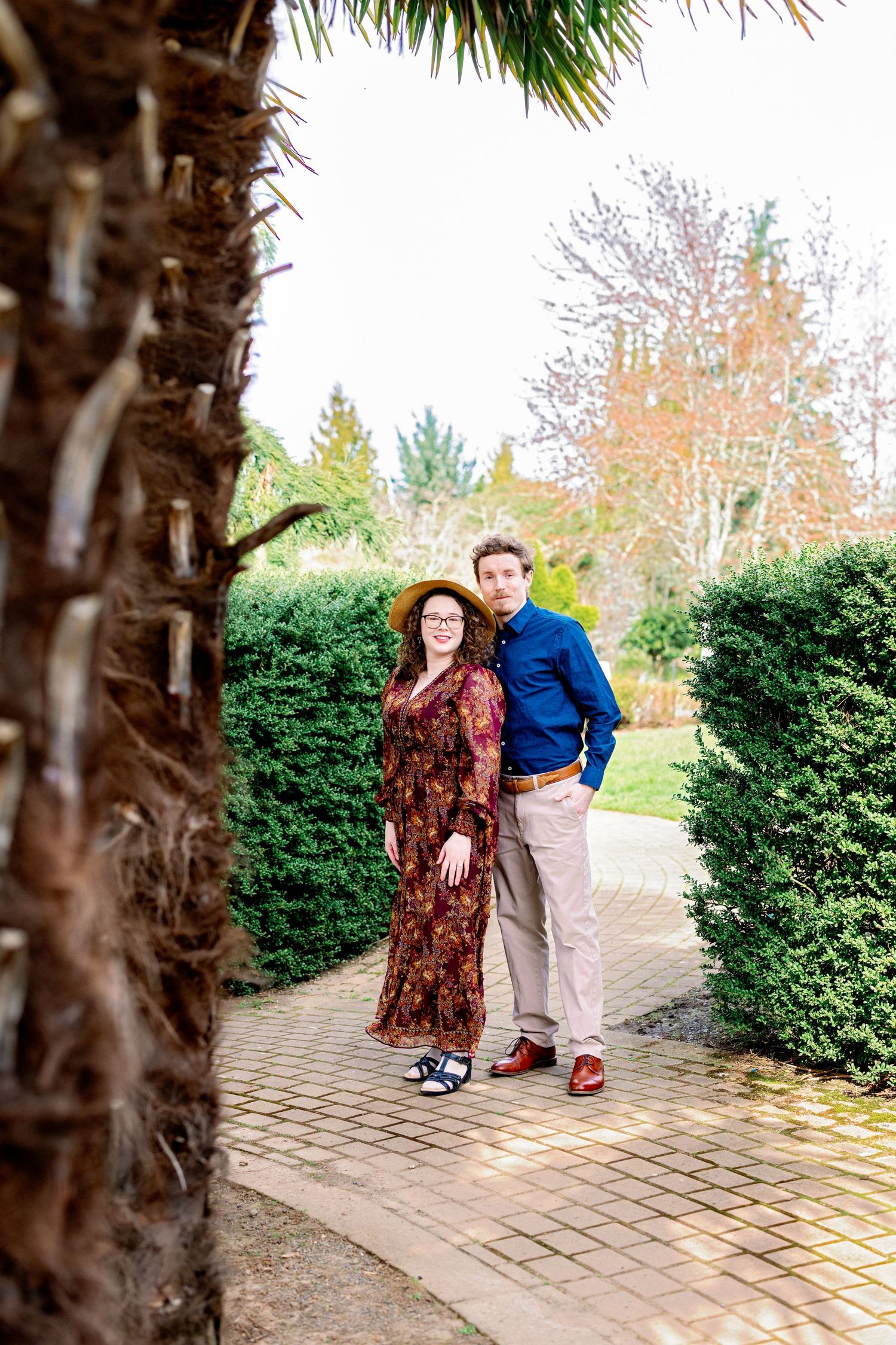 A couple shares a playful moment together on a brick pathway surrounded by green hedges and palm trees.
