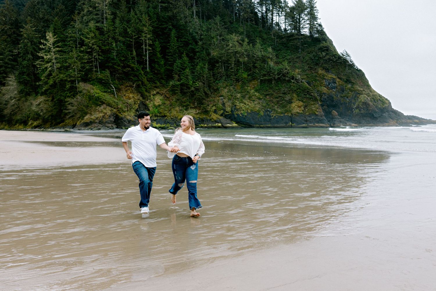Couple running hand-in-hand on a sandy beach with lush green forest cliffs in the background.