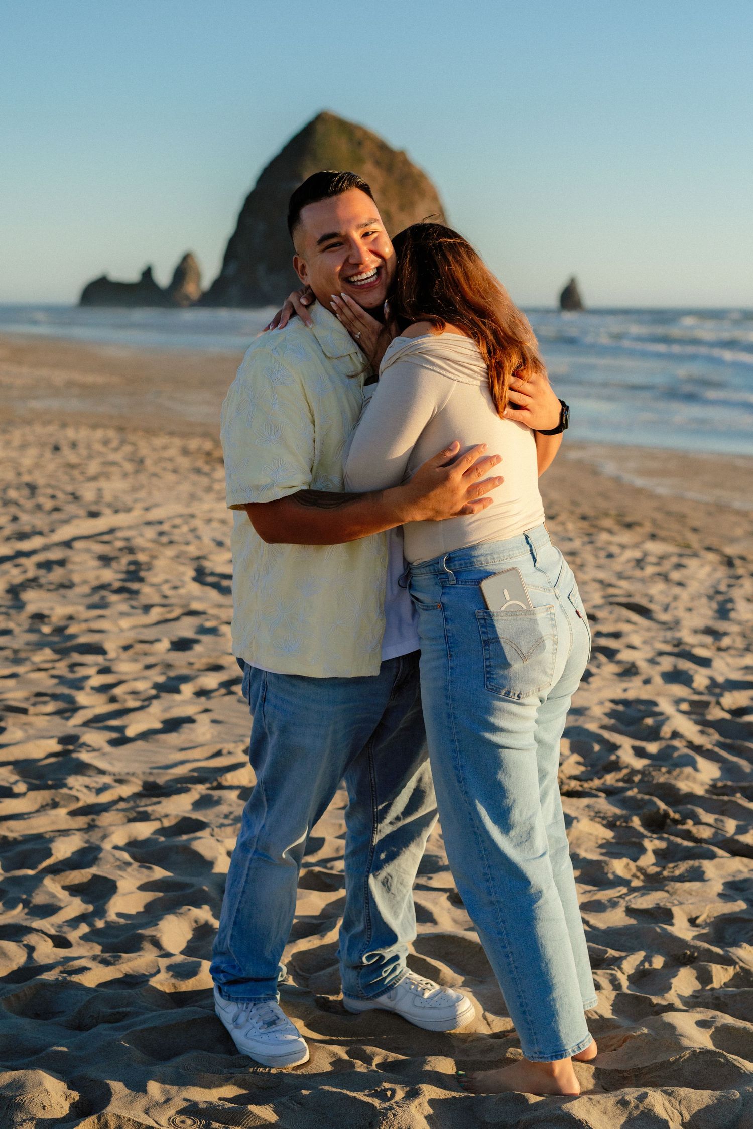 Happy couple embraces and laughs on sandy beach with Haystack Rock at Cannon Beach, Oregon during golden hour.