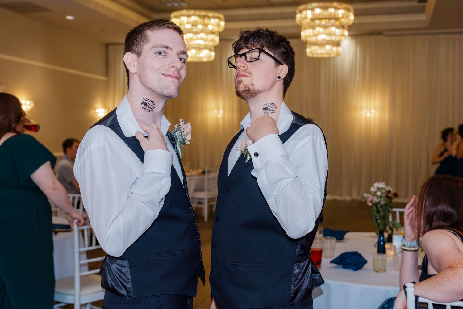 Two groomsmen in navy vests playfully pose pointing to their neck tattoos at a wedding reception.