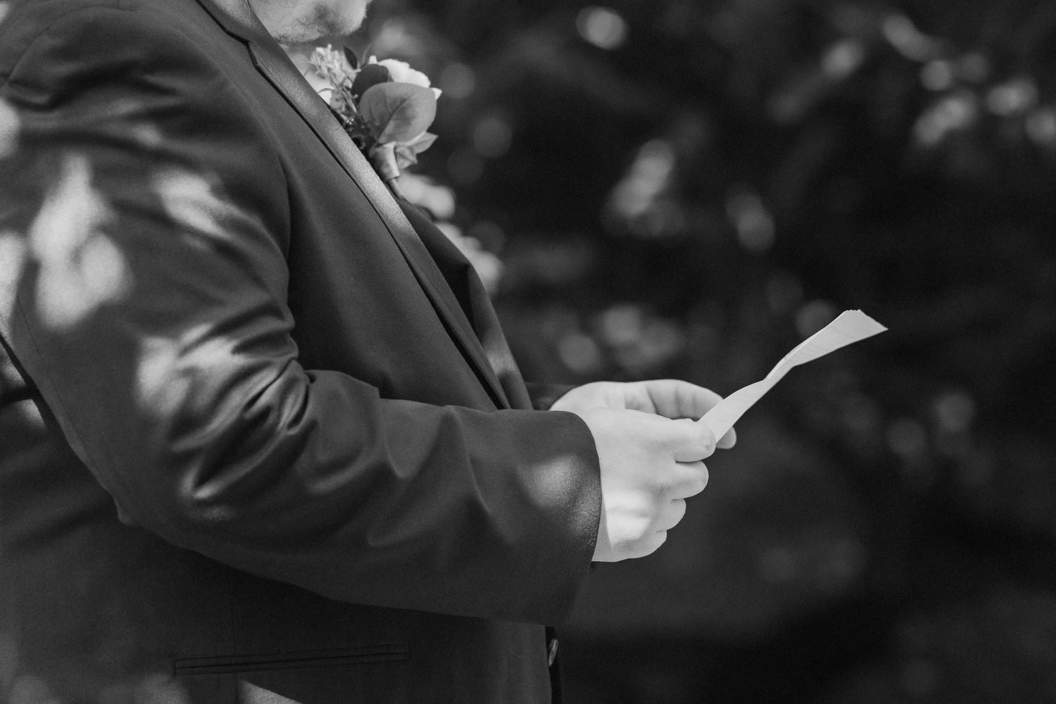 Groom in suit with boutonniere reading wedding vows from paper card outdoors in black and white photo.