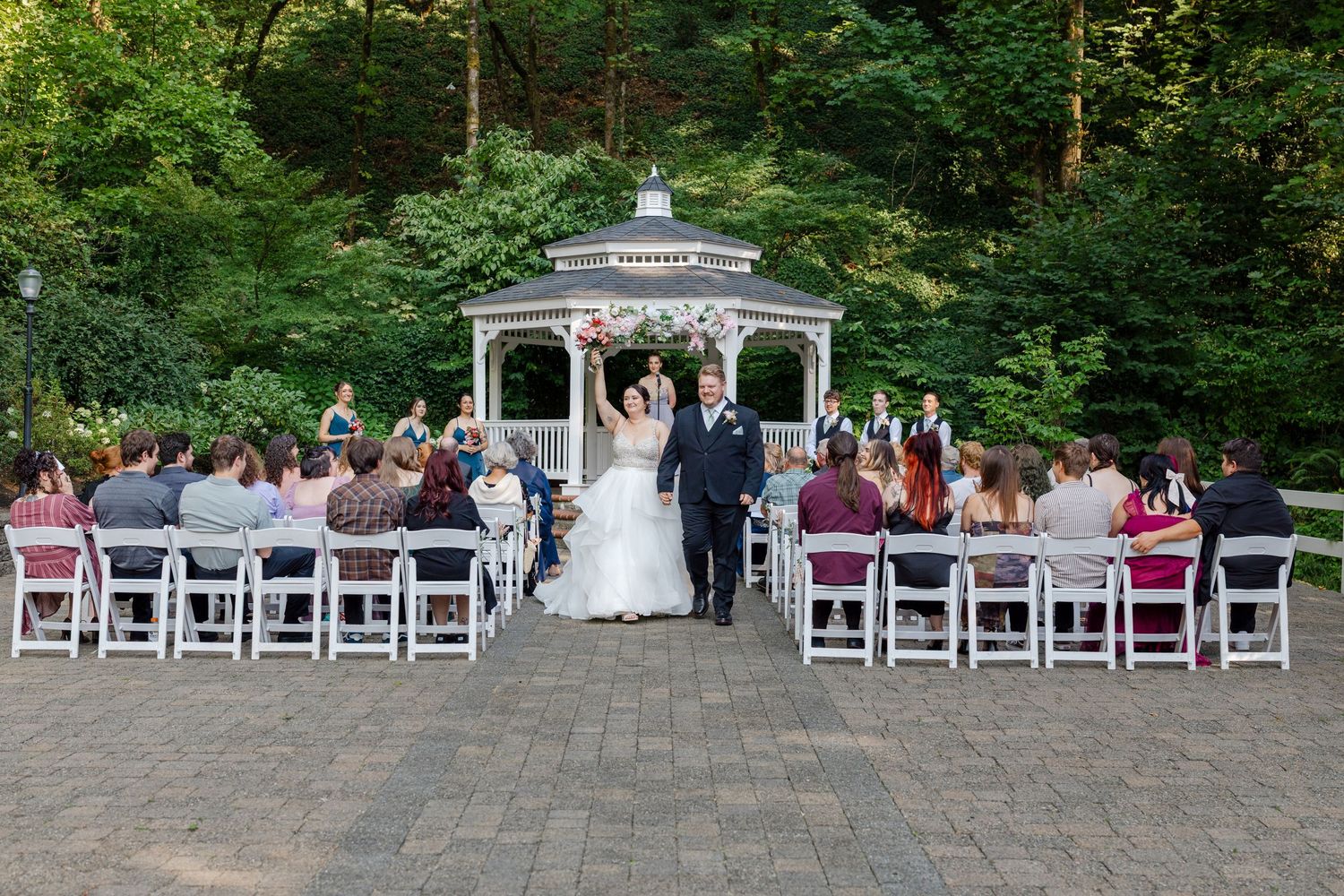 Newlyweds walk down the aisle after outdoor gazebo wedding ceremony with guests applauding.