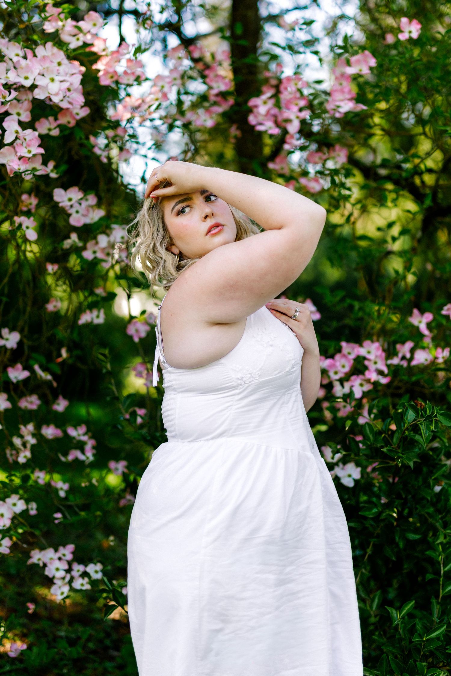 Woman in white dress posing among pink flowering trees in a lush green garden setting.