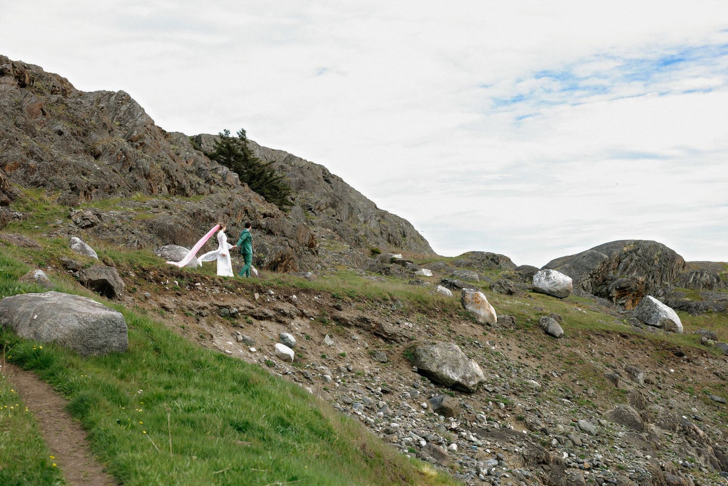 Hiker setting up a white tent on a rocky hillside trail with green grass and boulders under cloudy skies.