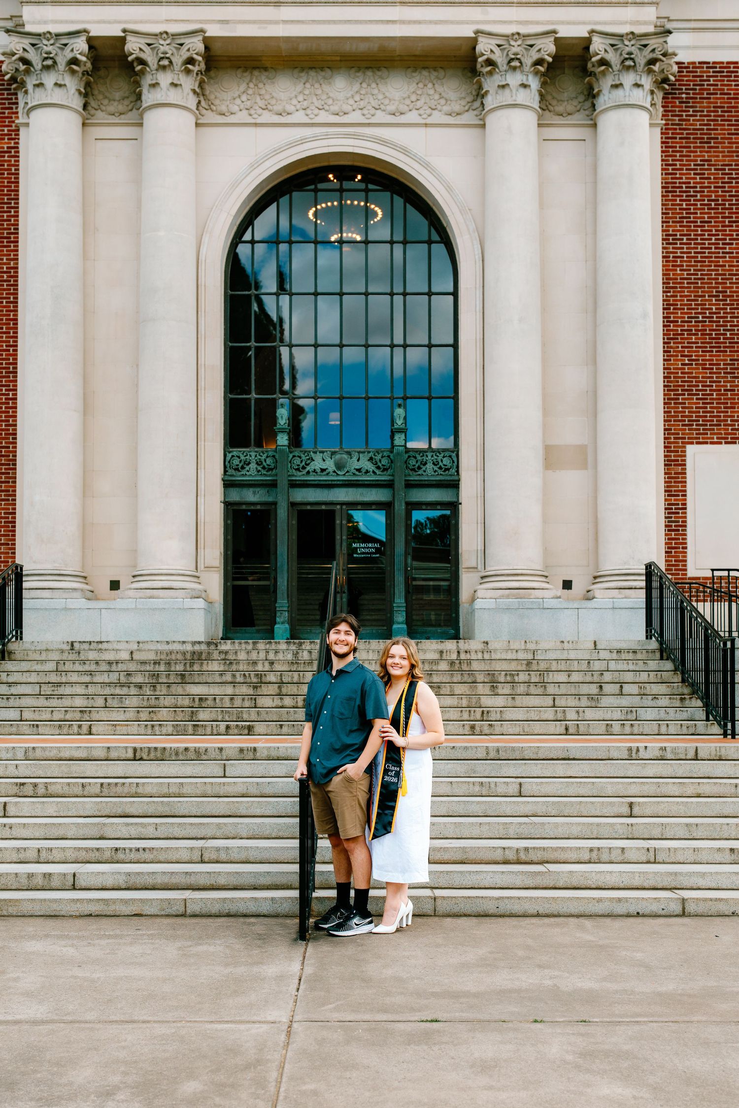Couple poses on steps of brick building with arched window and columns, woman wearing white dress and graduation stole.