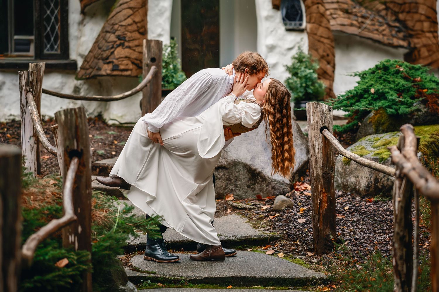 A romantic couple shares a passionate dip kiss in front of a rustic stone wall in an outdoor garden setting.