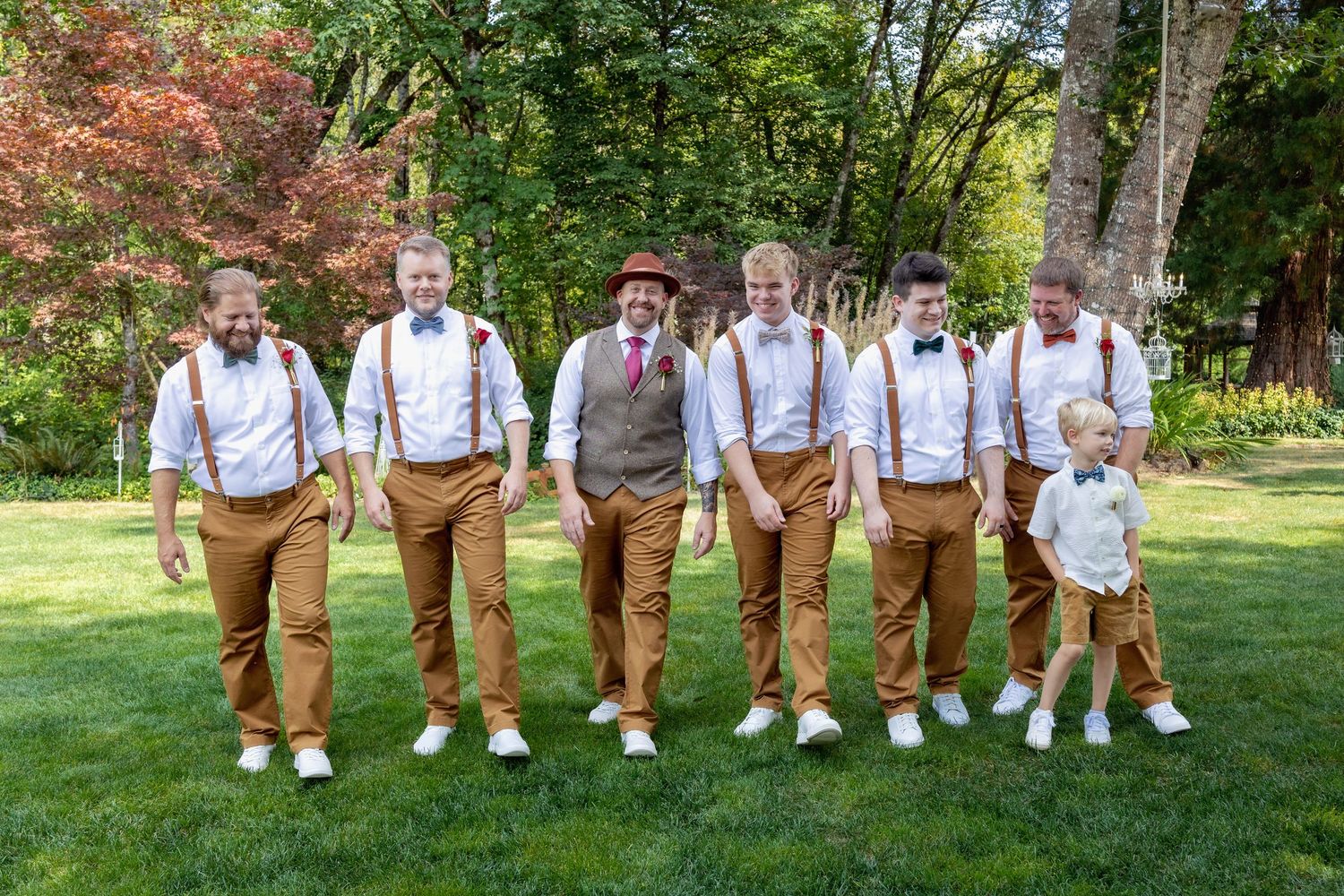 Groomsmen in matching brown pants and suspenders lined up on grass for wedding photo.