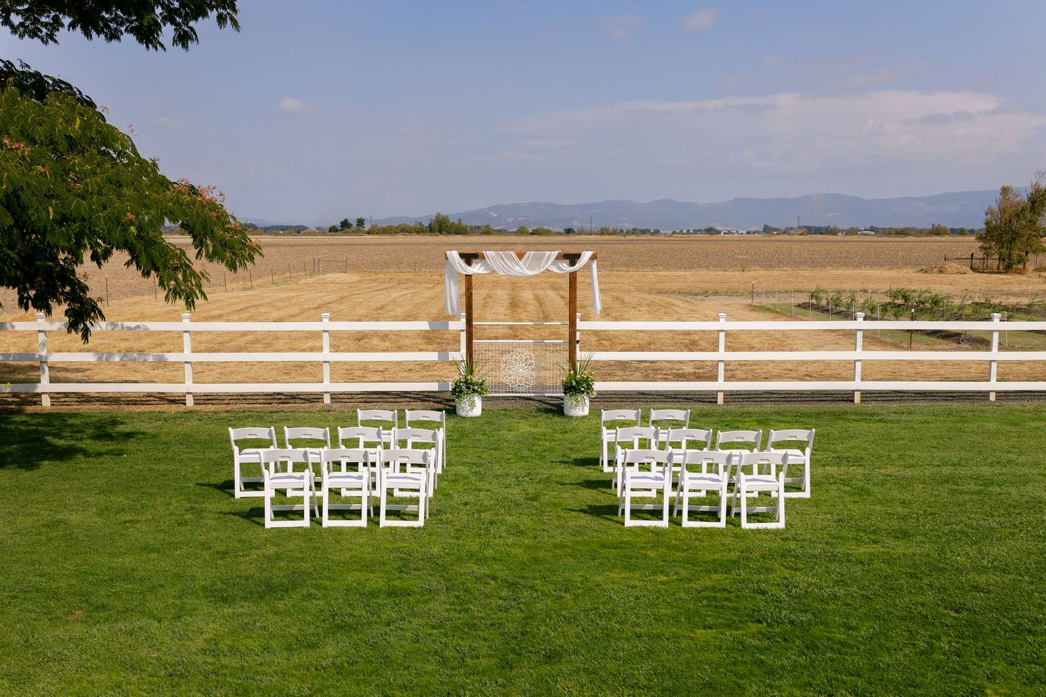 White wedding ceremony chairs arranged on a scenic lawn with white fencing and mountain views.